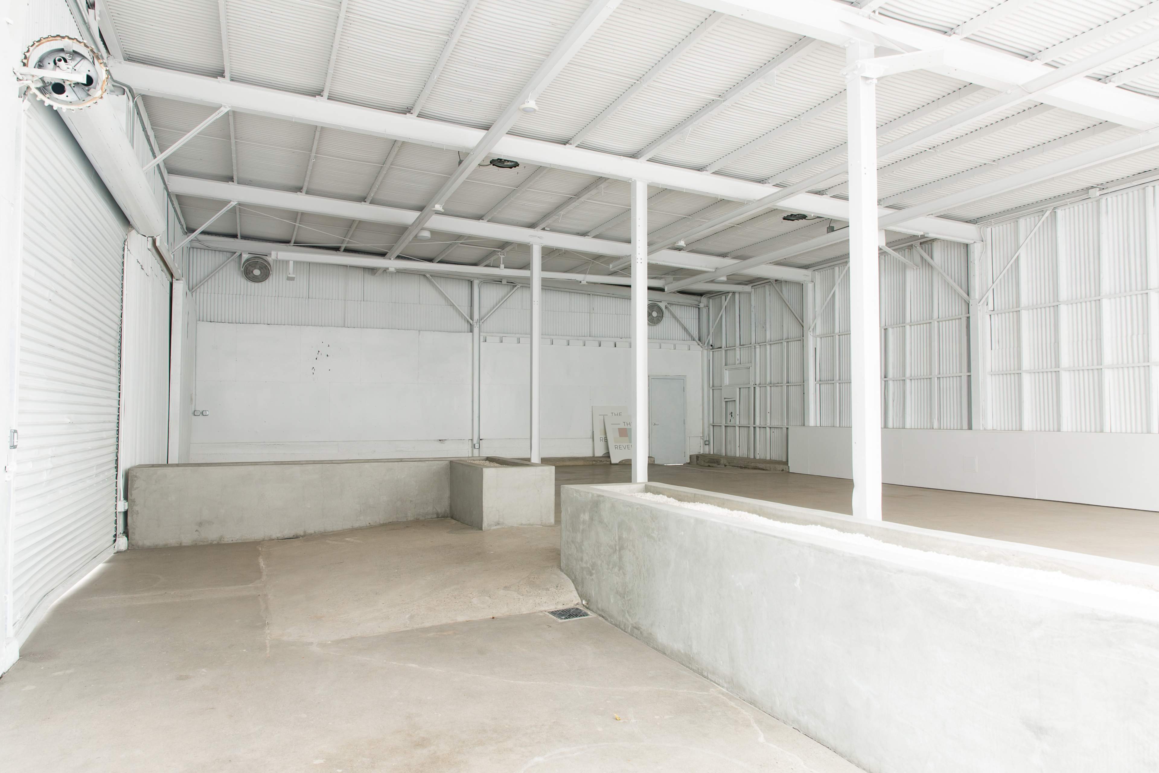 The image shows an empty industrial space with white walls, concrete flooring, and large overhead beams.
