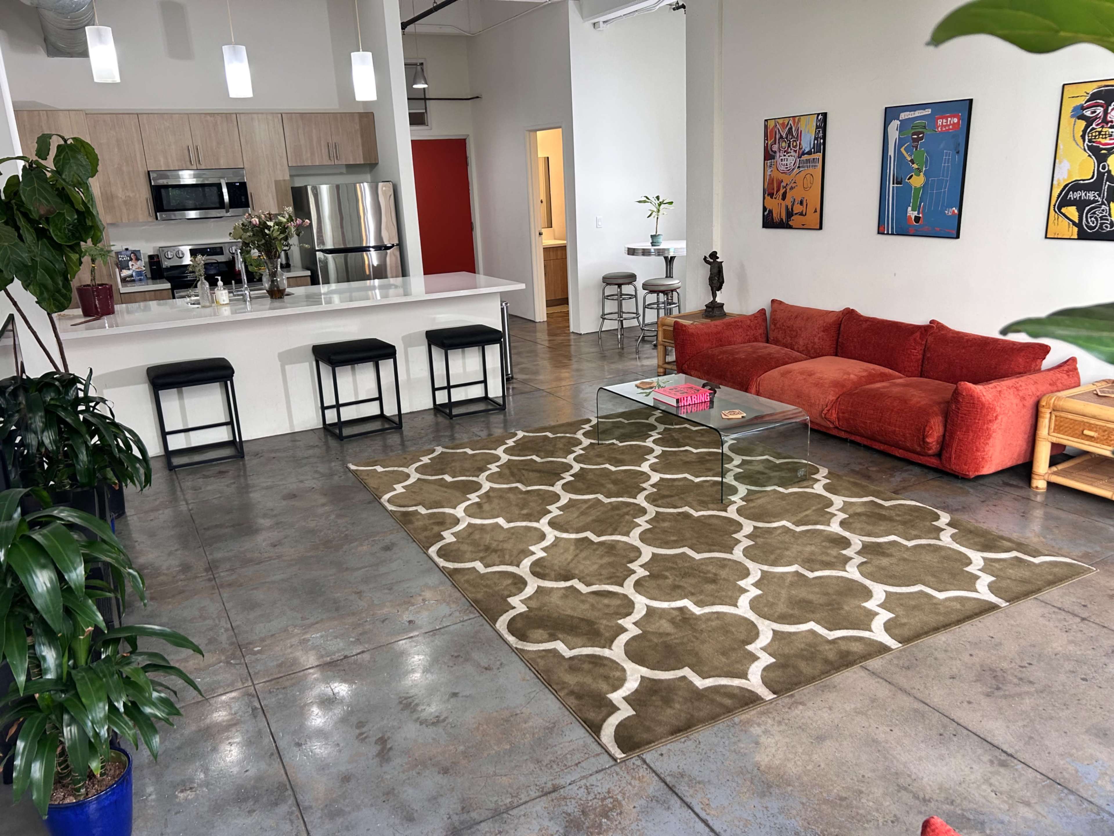 The image shows a spacious, modern living area with a red sofa, a large patterned rug, and a kitchen area featuring stainless steel appliances and a bar with black stools.