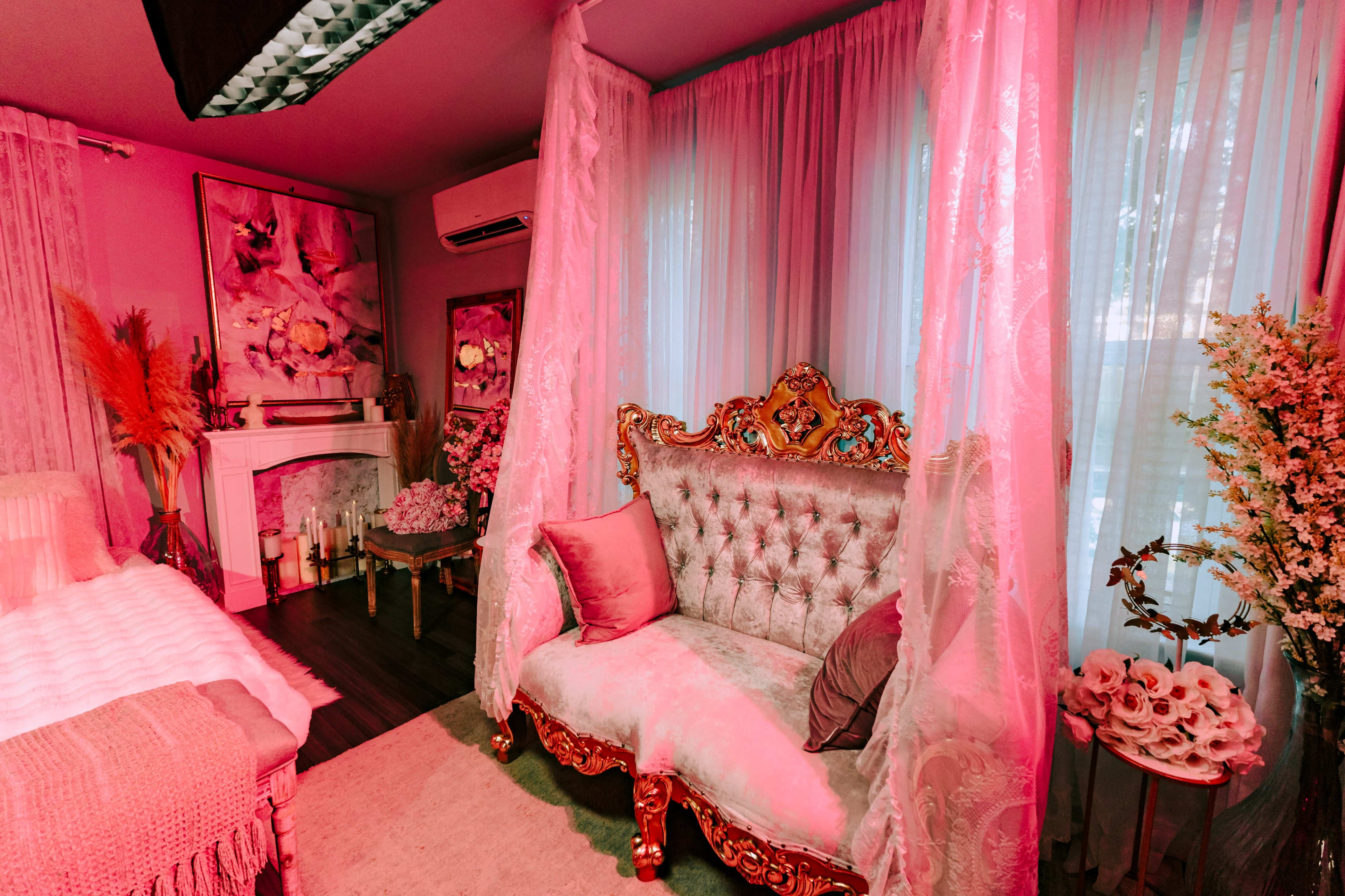 The image shows a room decorated with ornate furniture, featuring a plush vintage-style sofa and floral arrangements, draped with sheer curtains and illuminated in soft pink light.