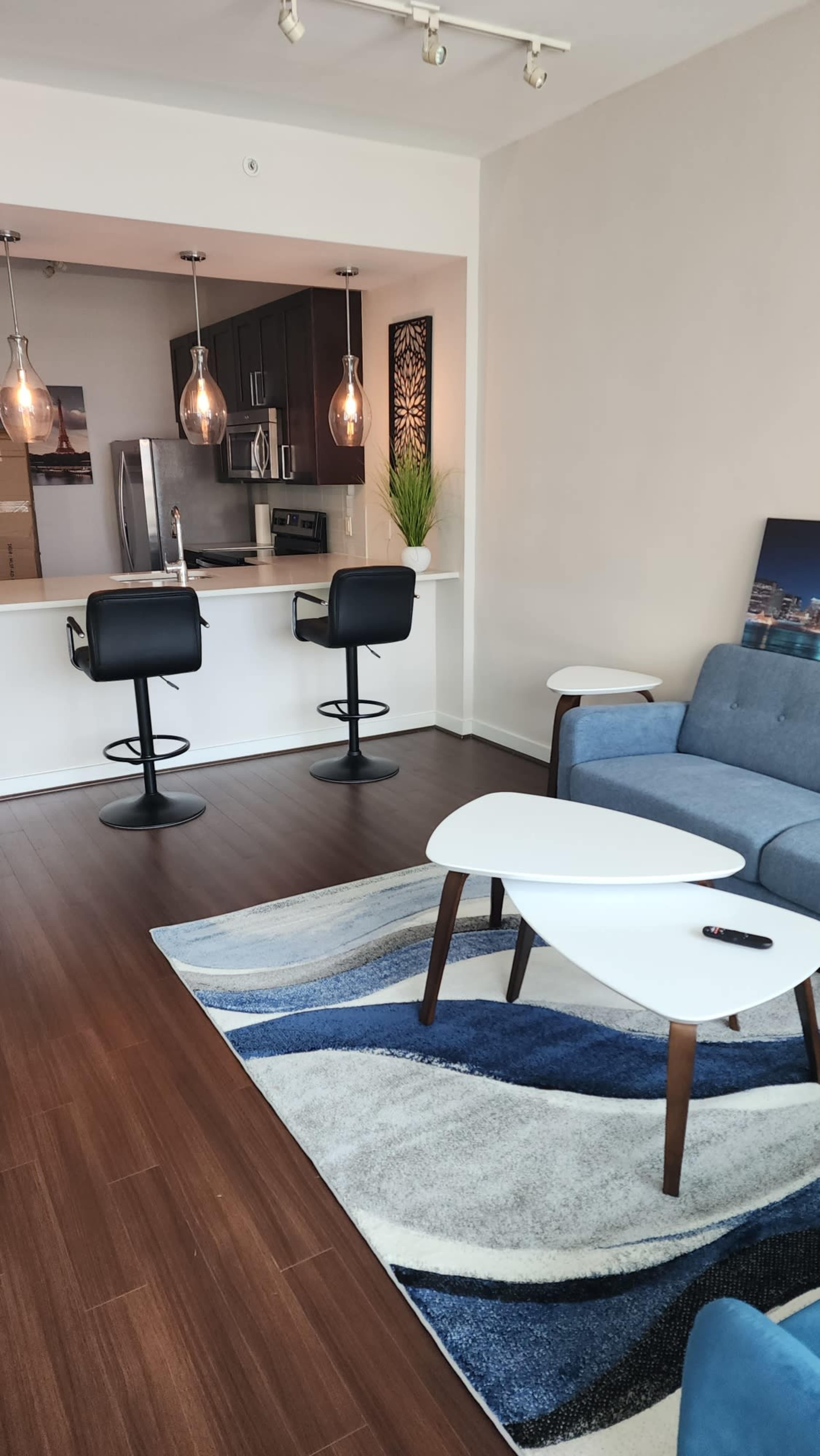 The scene shows a modern living area featuring a blue sofa, a coffee table with an abstract rug, and a kitchen with bar stools visible in the background.
