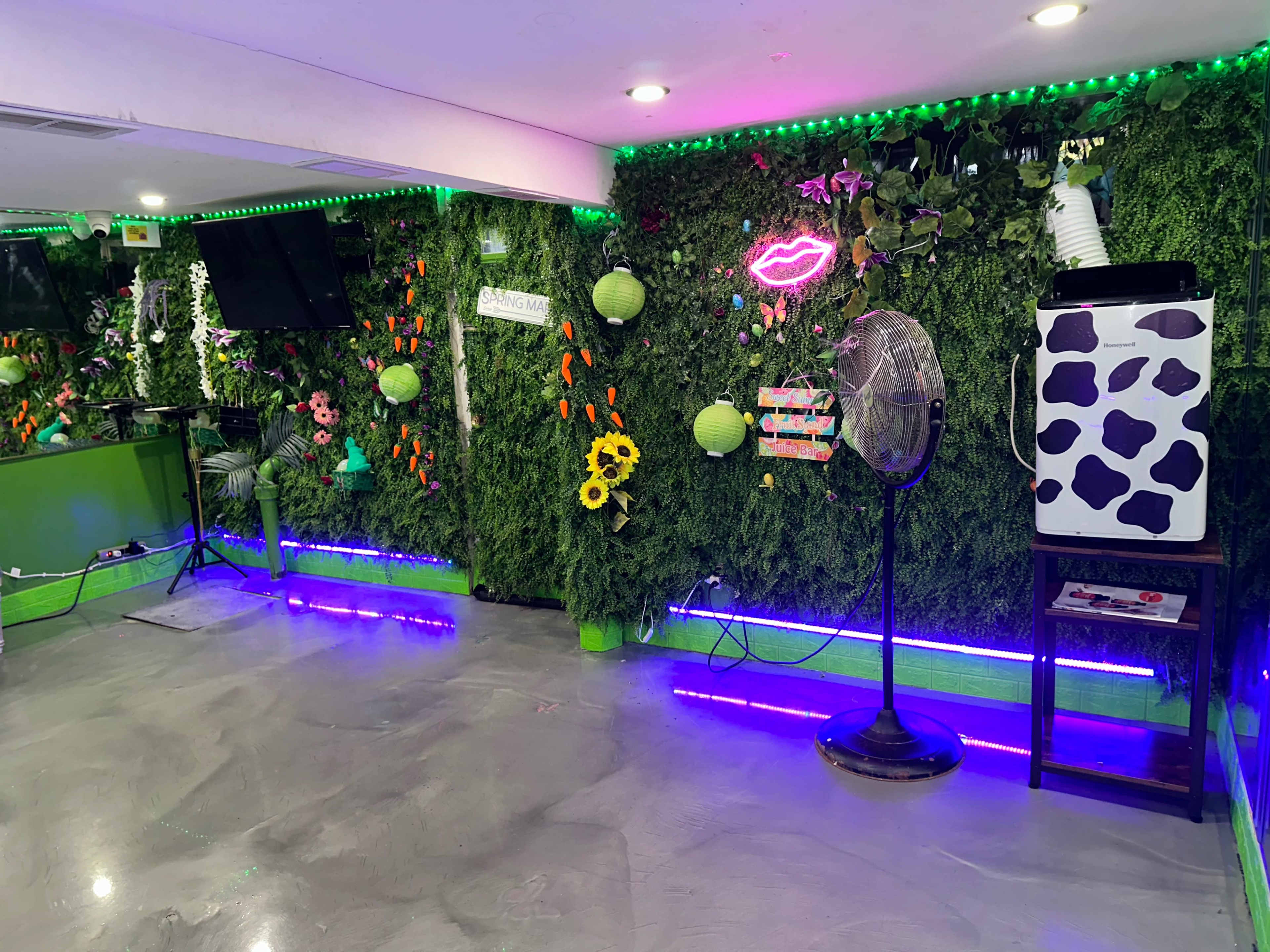 A vibrant indoor space with a green wall adorned with plants, neon signs, and decorative elements, featuring a fan and a whimsical, cow-patterned ice cream machine.