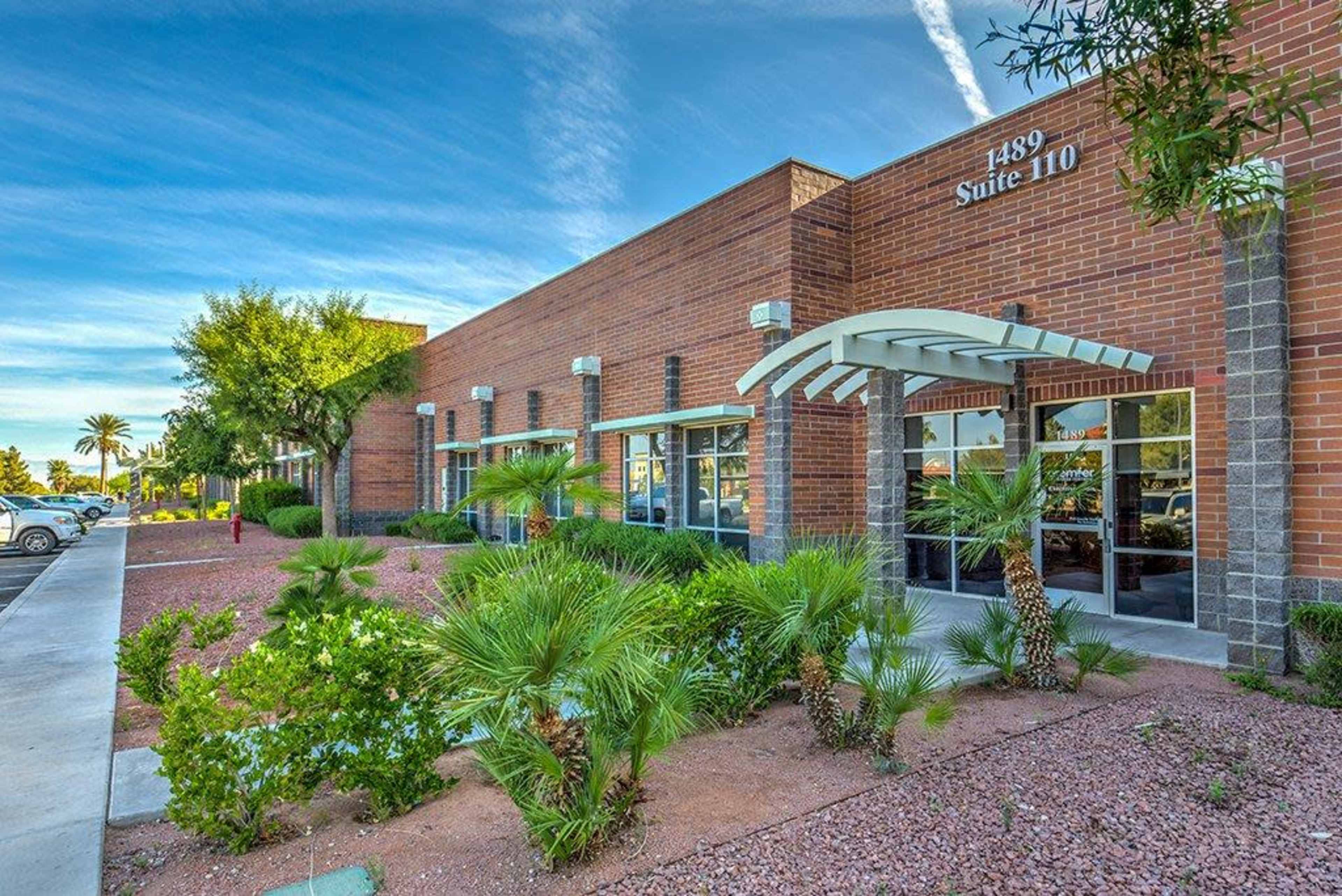 The image shows a brick commercial building with a landscaped front featuring small palm trees and shrubs.