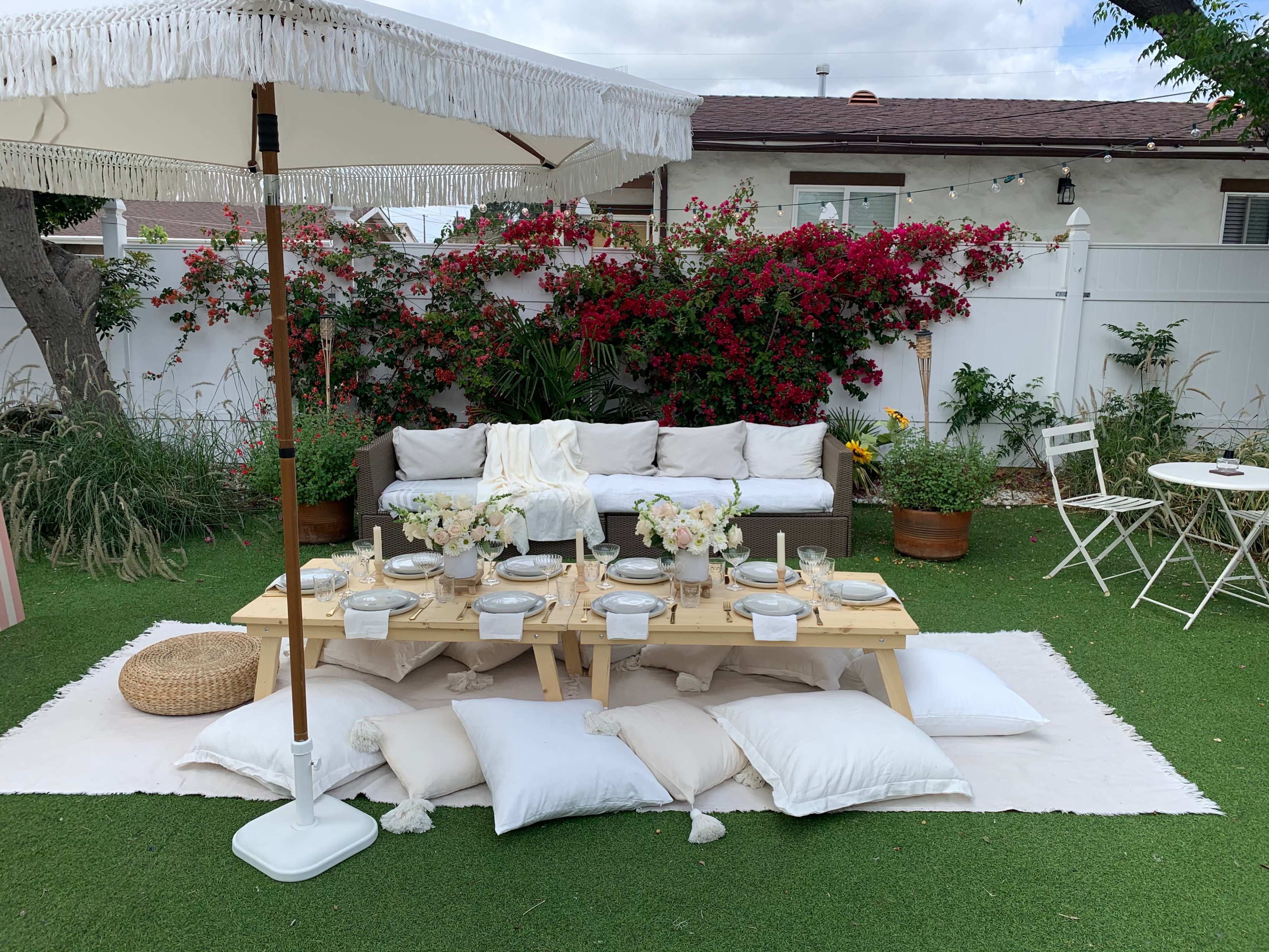 A cozy outdoor setup featuring a low wooden table with decorative plates and centerpieces, surrounded by a white couch, pillows, and a large umbrella, against a backdrop of blooming bougainvillea.