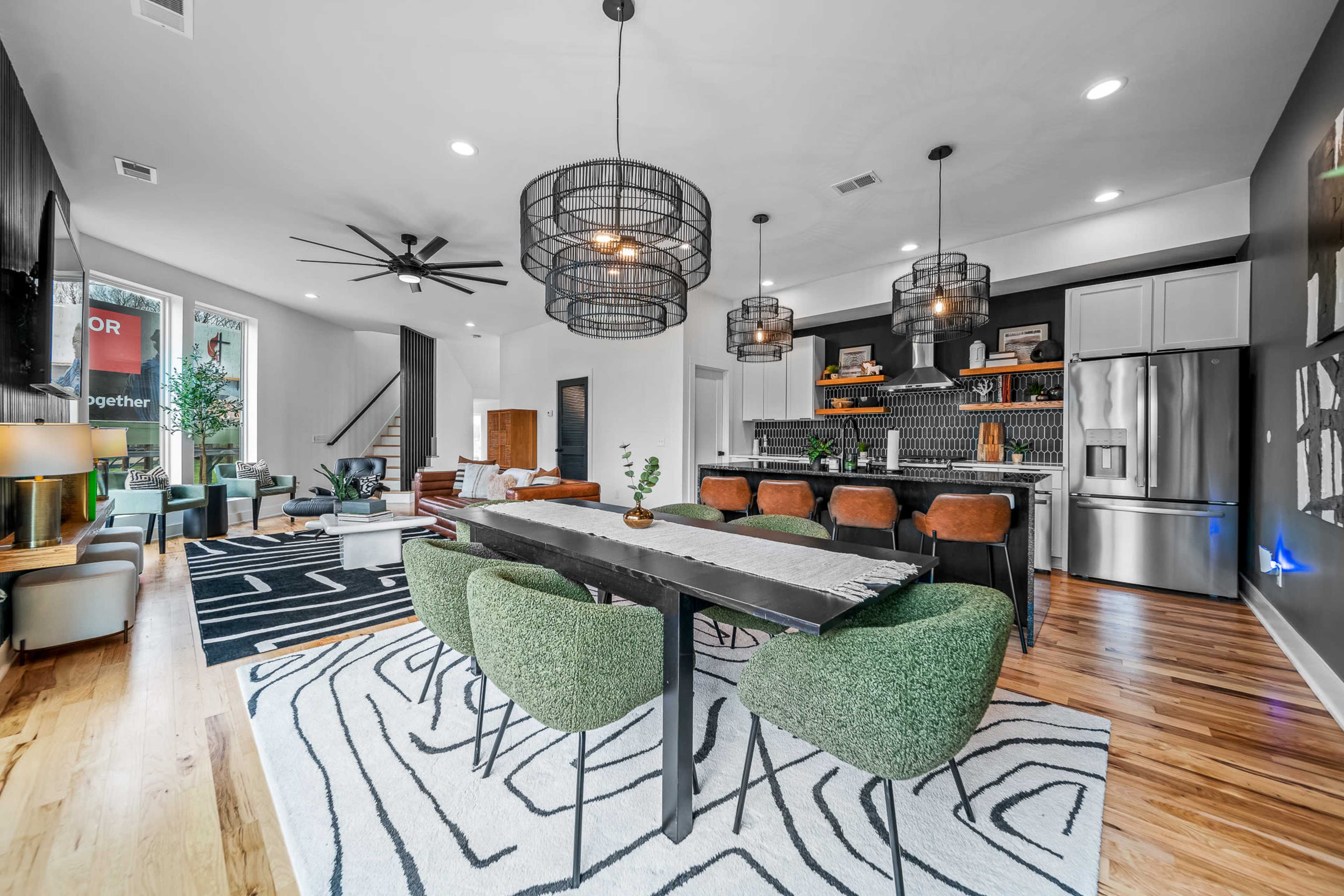 A modern open-concept living and dining area featuring a large dining table with green chairs, pendant lights, and a stylish kitchen with stainless steel appliances.