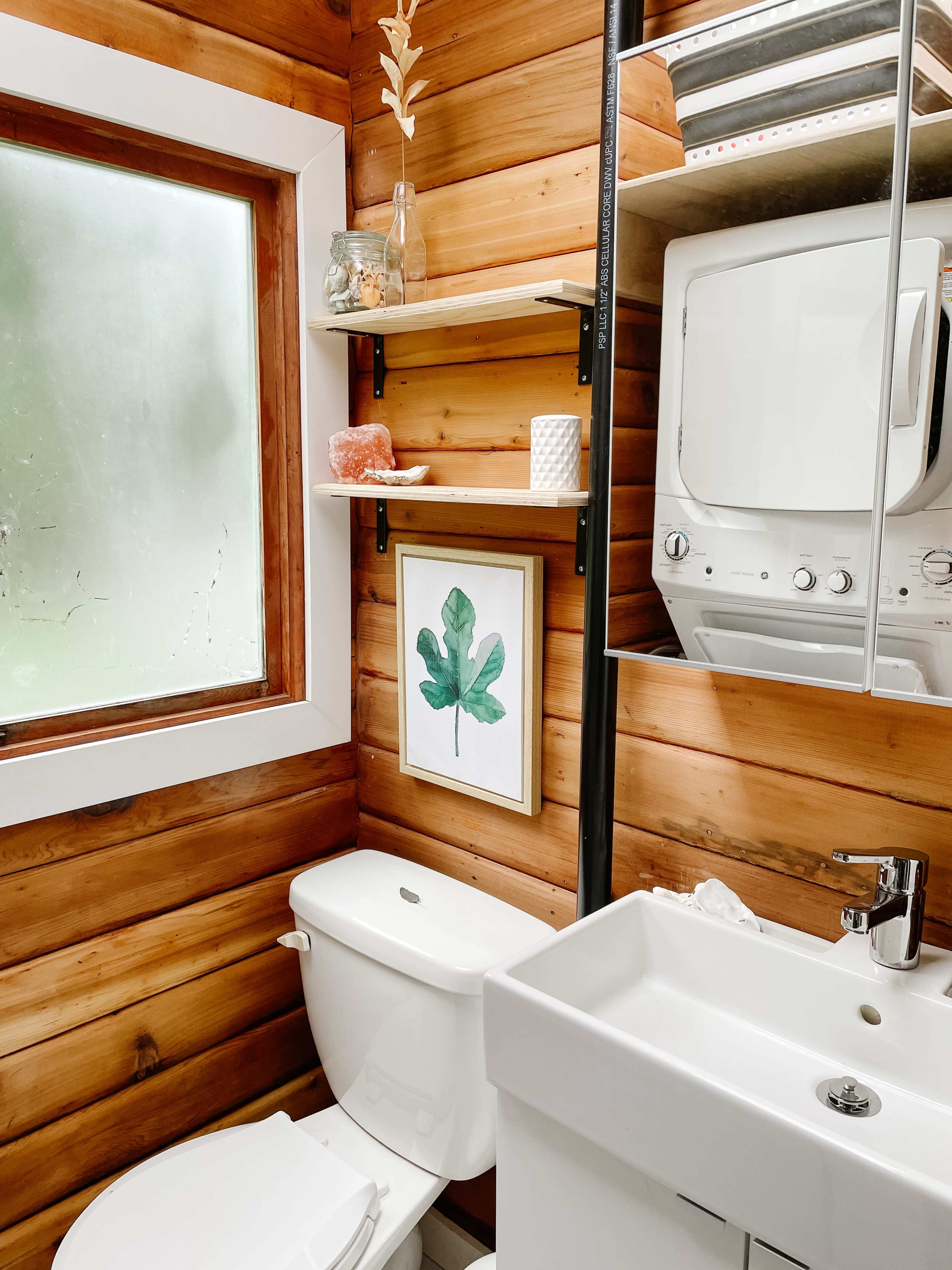 A small bathroom features wooden walls, a wall-mounted sink, a modern toilet, and shelves with decorative items and a framed leaf print.
