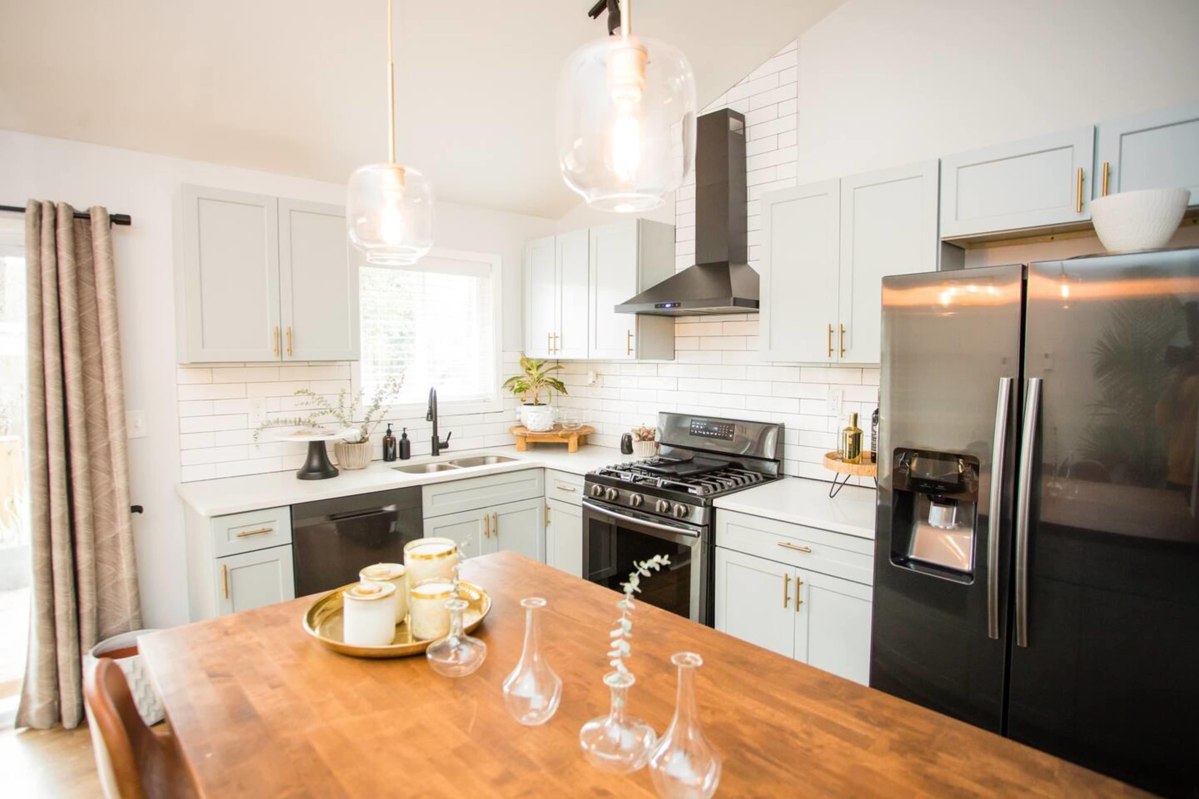 The image shows a modern kitchen with light blue cabinets, a black refrigerator, a gas stove, and a wooden dining table with decorative items.