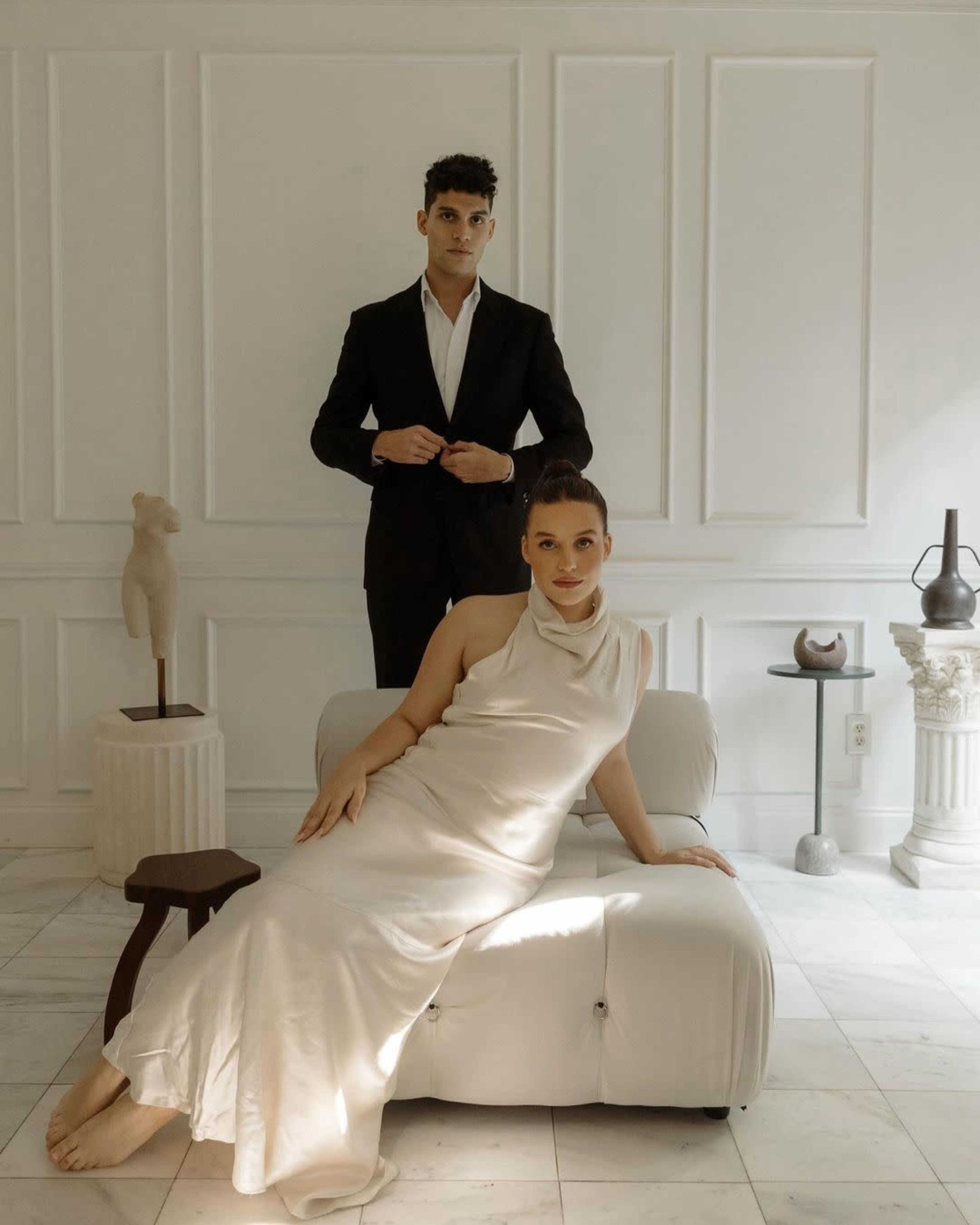 A man in a black suit stands behind a woman in a white dress, who is seated on a light-colored chaise lounge in a well-lit, minimalist room.