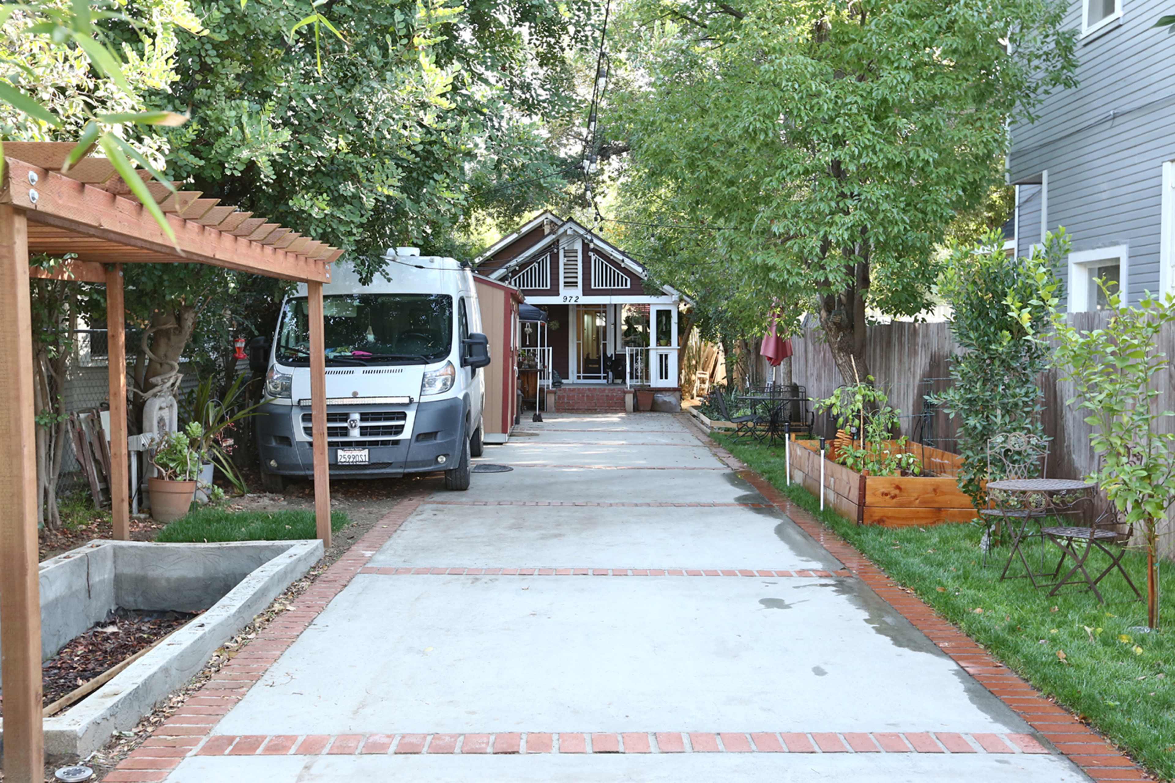 The image shows a narrow driveway lined with brick, leading to a house surrounded by trees and garden features.
