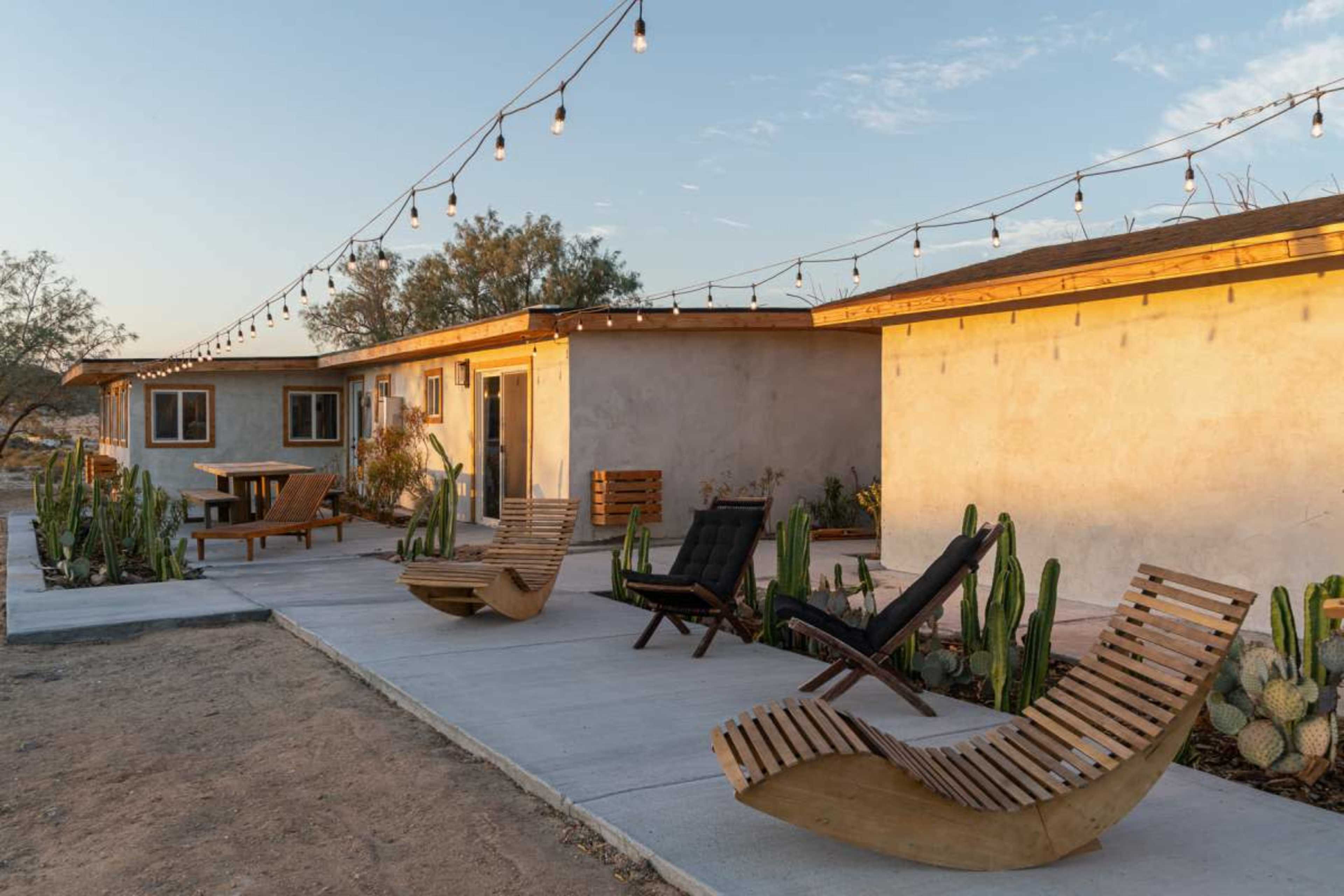 The image depicts a desert landscape featuring two modern, stucco buildings with outdoor seating, illuminated by string lights above.