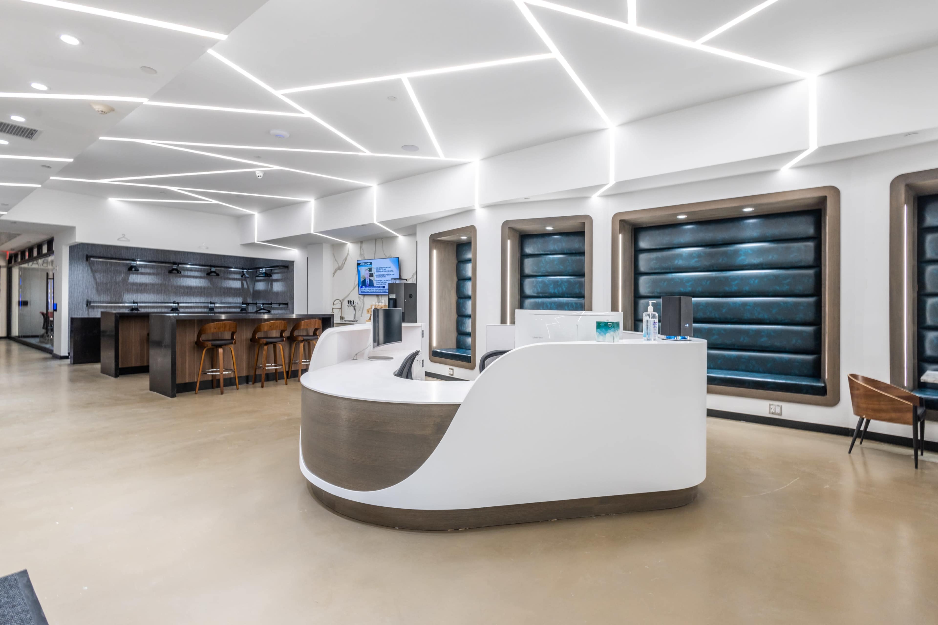 A modern reception area with a curved white desk, brown bar stools, and blue upholstered panels along the walls, illuminated by geometric ceiling lights.