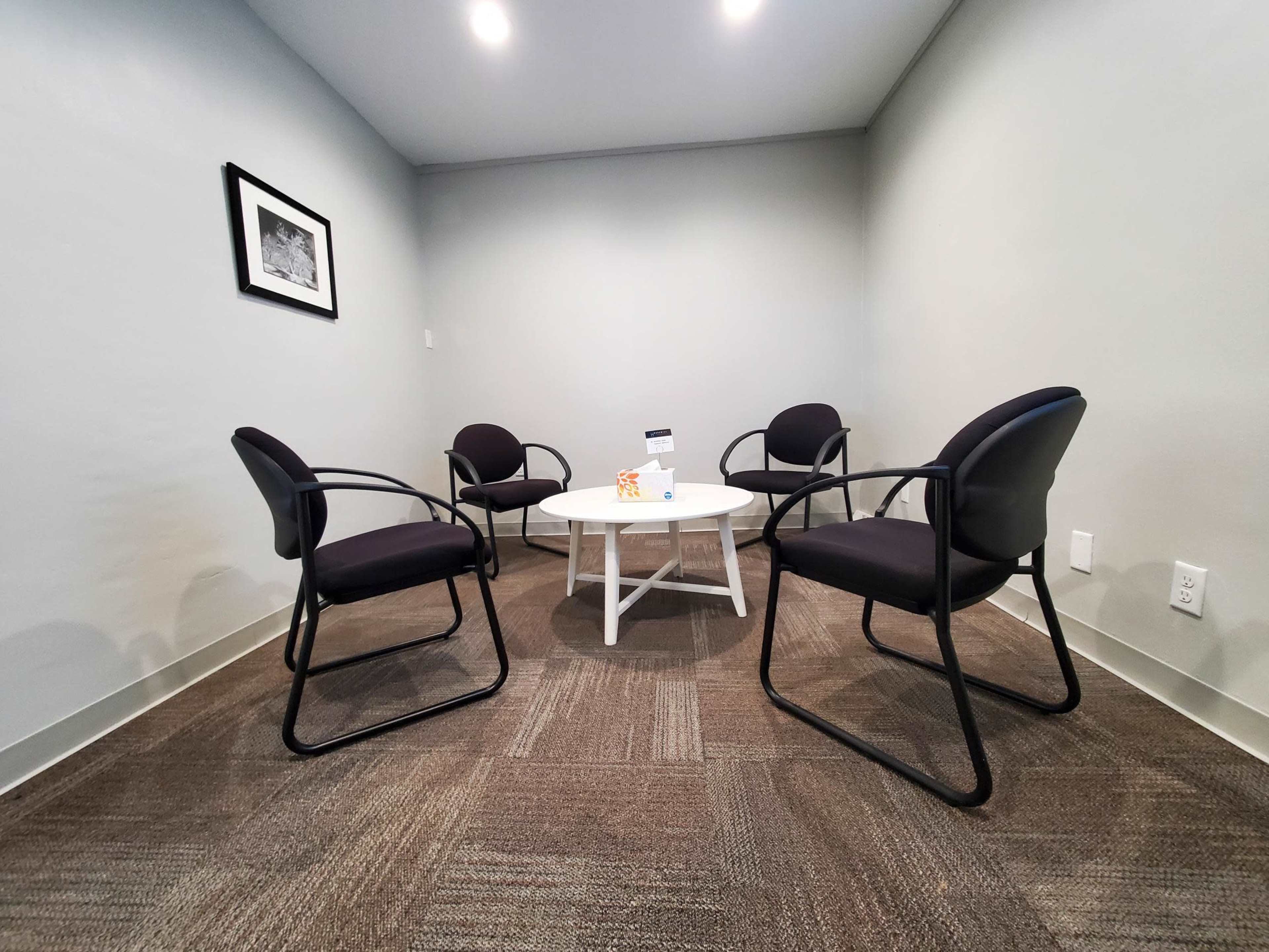 A small meeting room with four black chairs arranged around a white table, set against a gray wall.