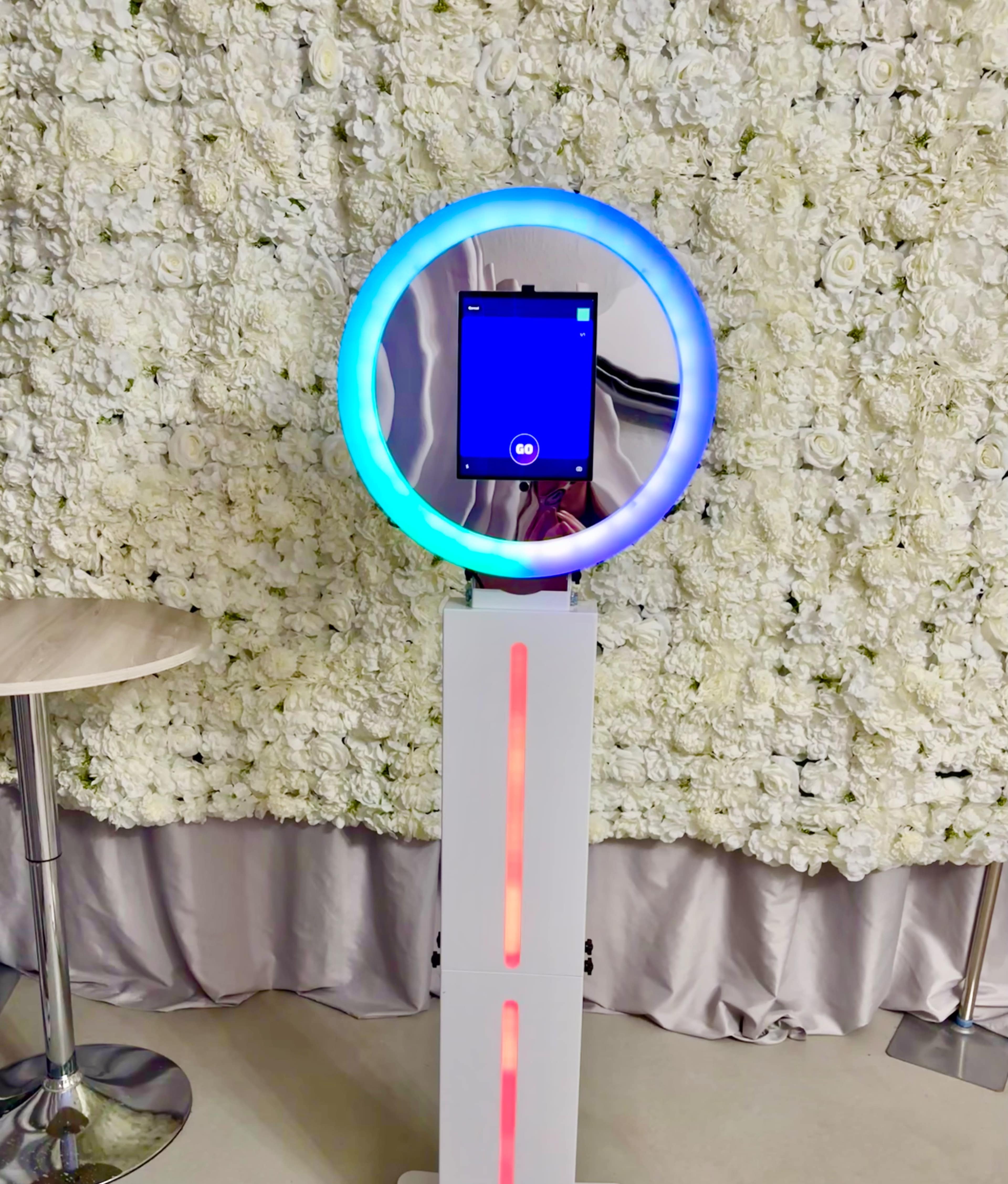 A photo booth with a circular LED light ring stands in front of a wall covered in white flowers.