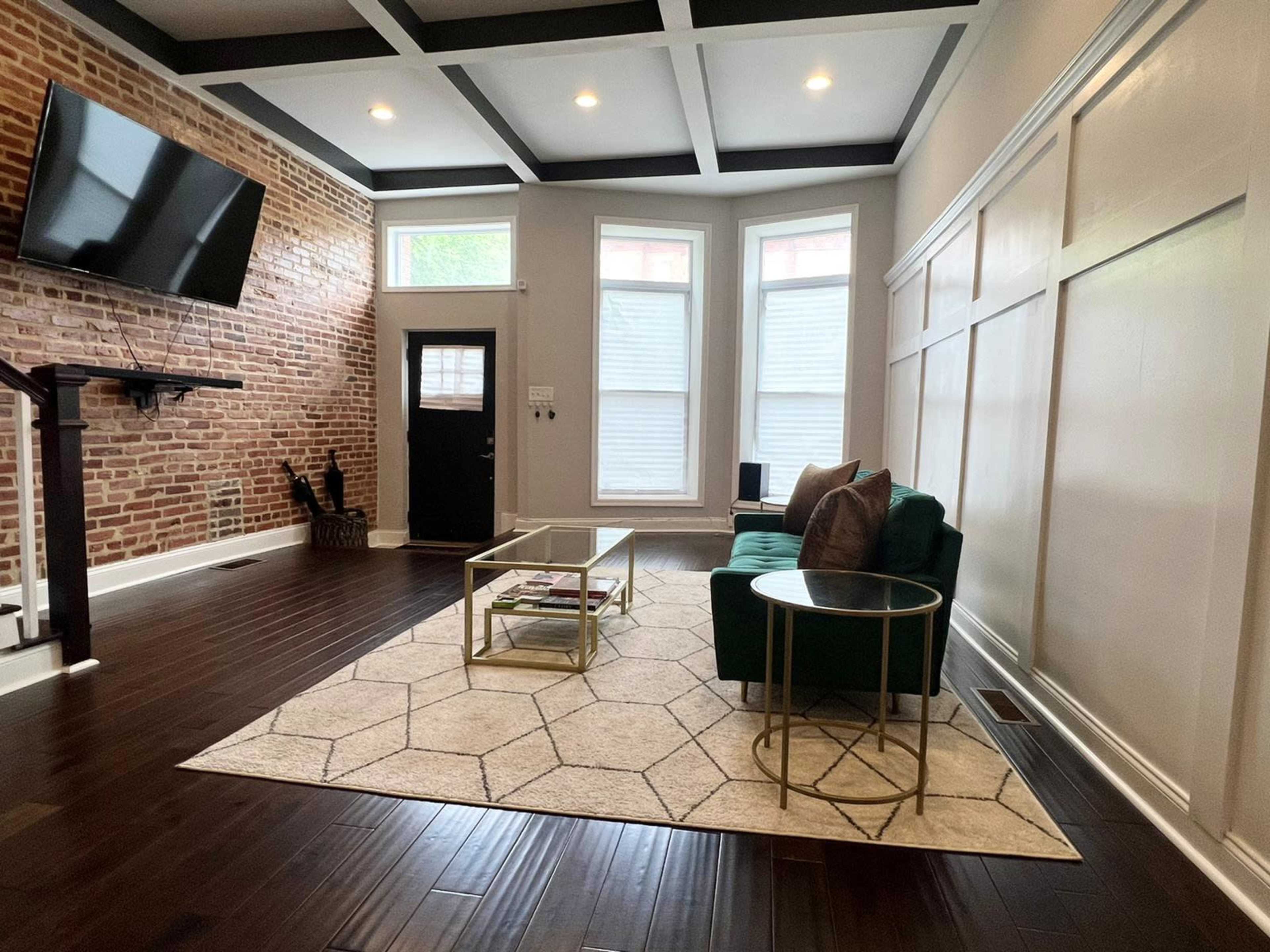 A modern living room with a green couch, a gold coffee table, and a brick wall, featuring large windows and a dark door.