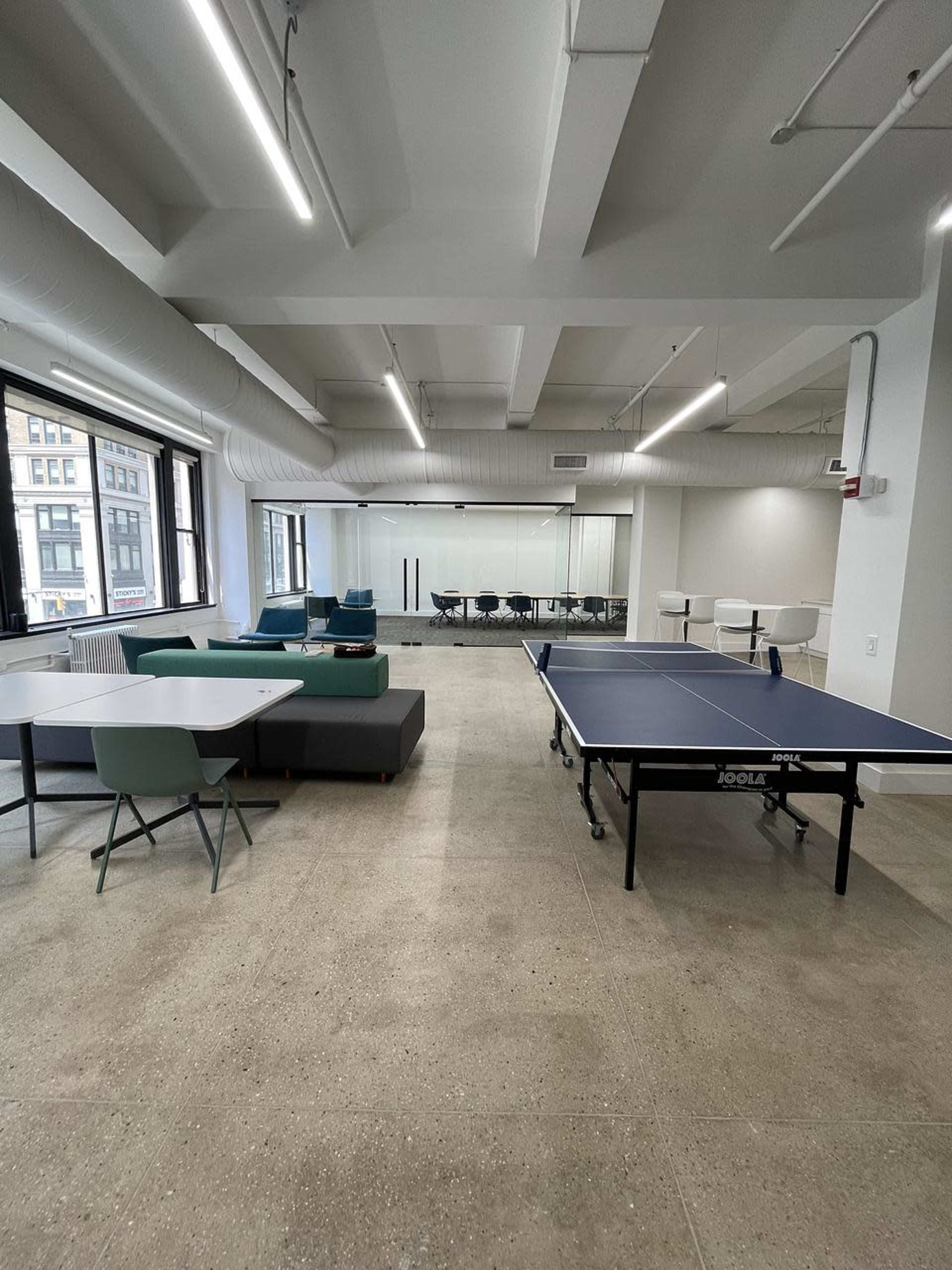 The image shows a modern office space featuring a ping pong table, lounge seating, and a glass-walled conference room.