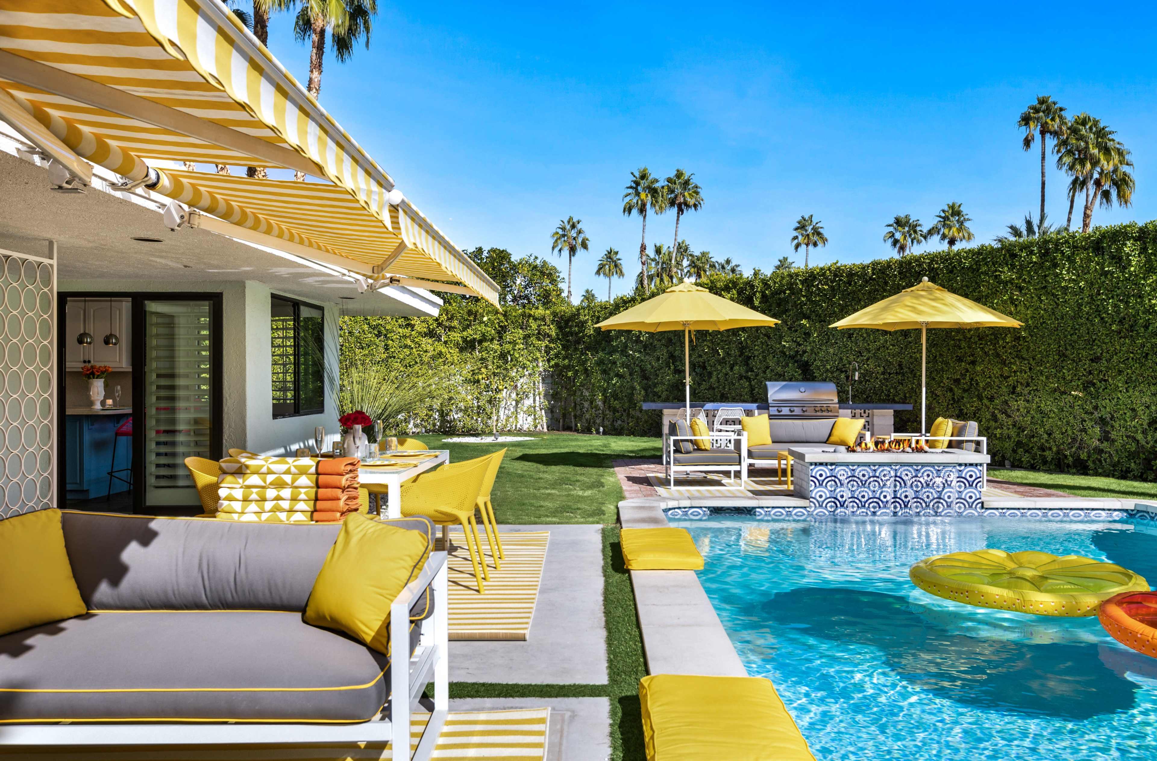 The scene features a vibrant outdoor patio area with yellow accents, a swimming pool, and a barbecue setup surrounded by palm trees.