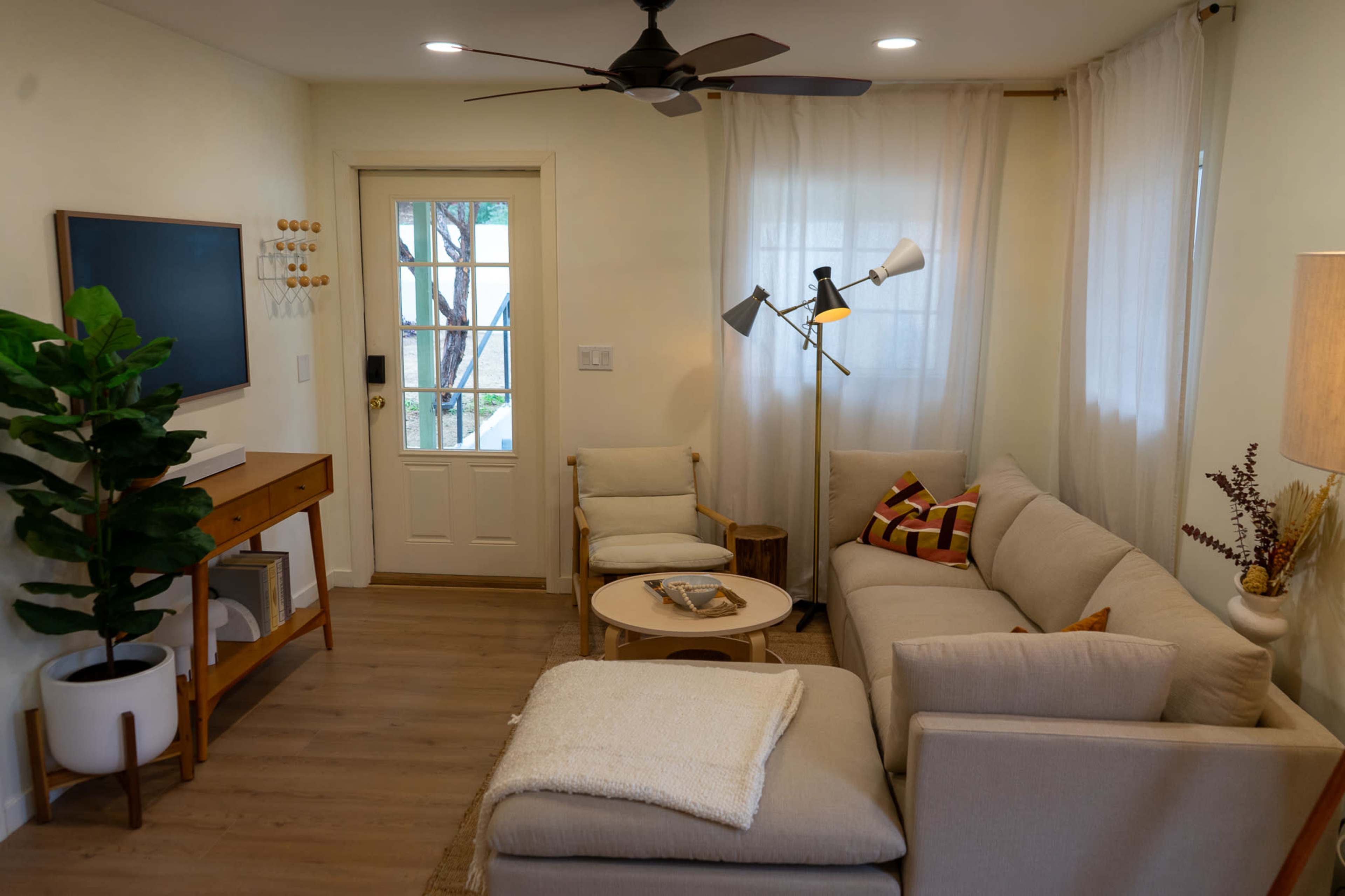 The image shows a cozy living room with a light-colored sofa, a small round coffee table, and a door leading to the outside, alongside a potted plant and modern lighting fixtures.