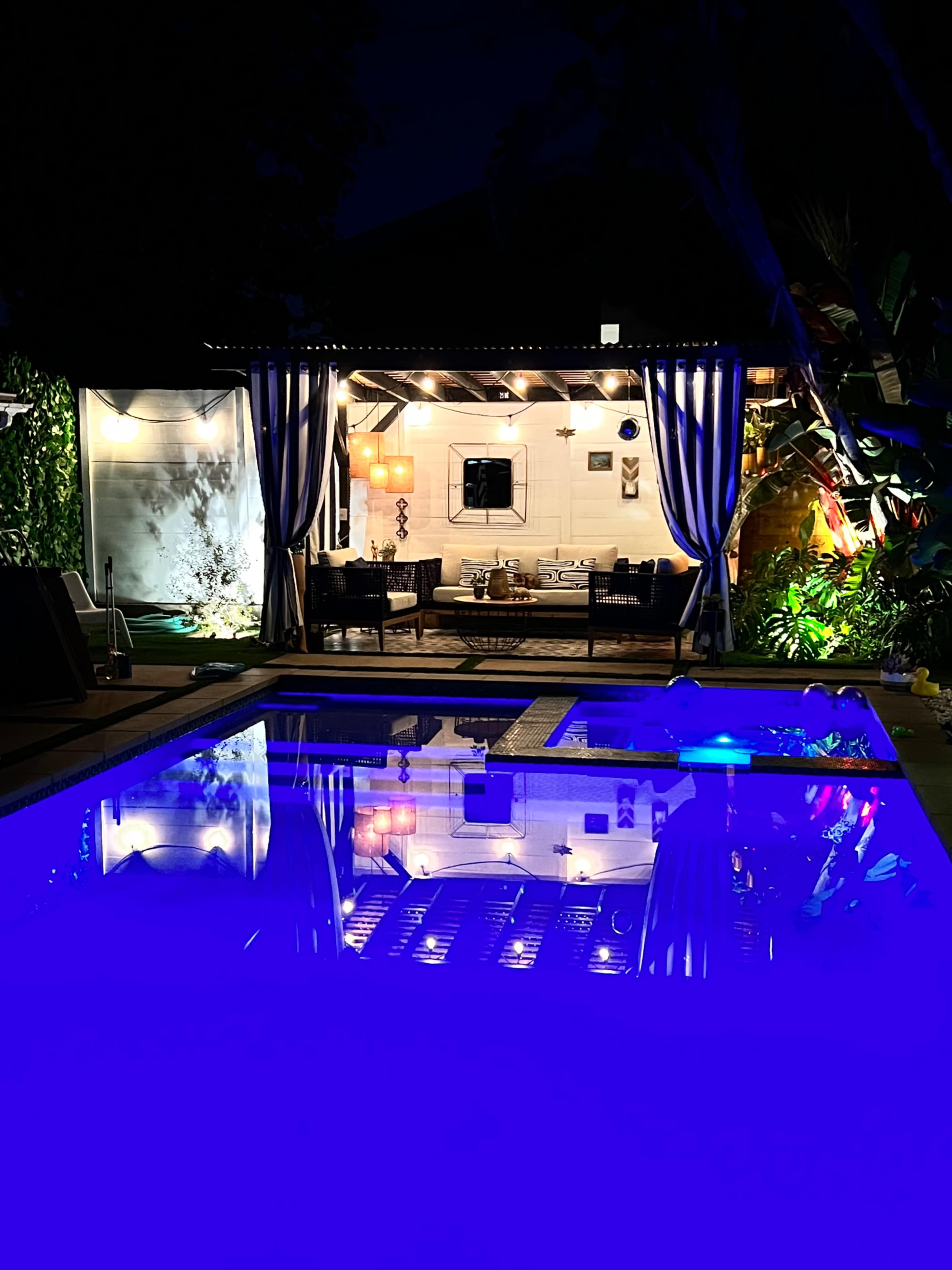 A well-lit outdoor lounge area with seating is reflected in a pool at night, surrounded by greenery and illuminated by ambient lighting.