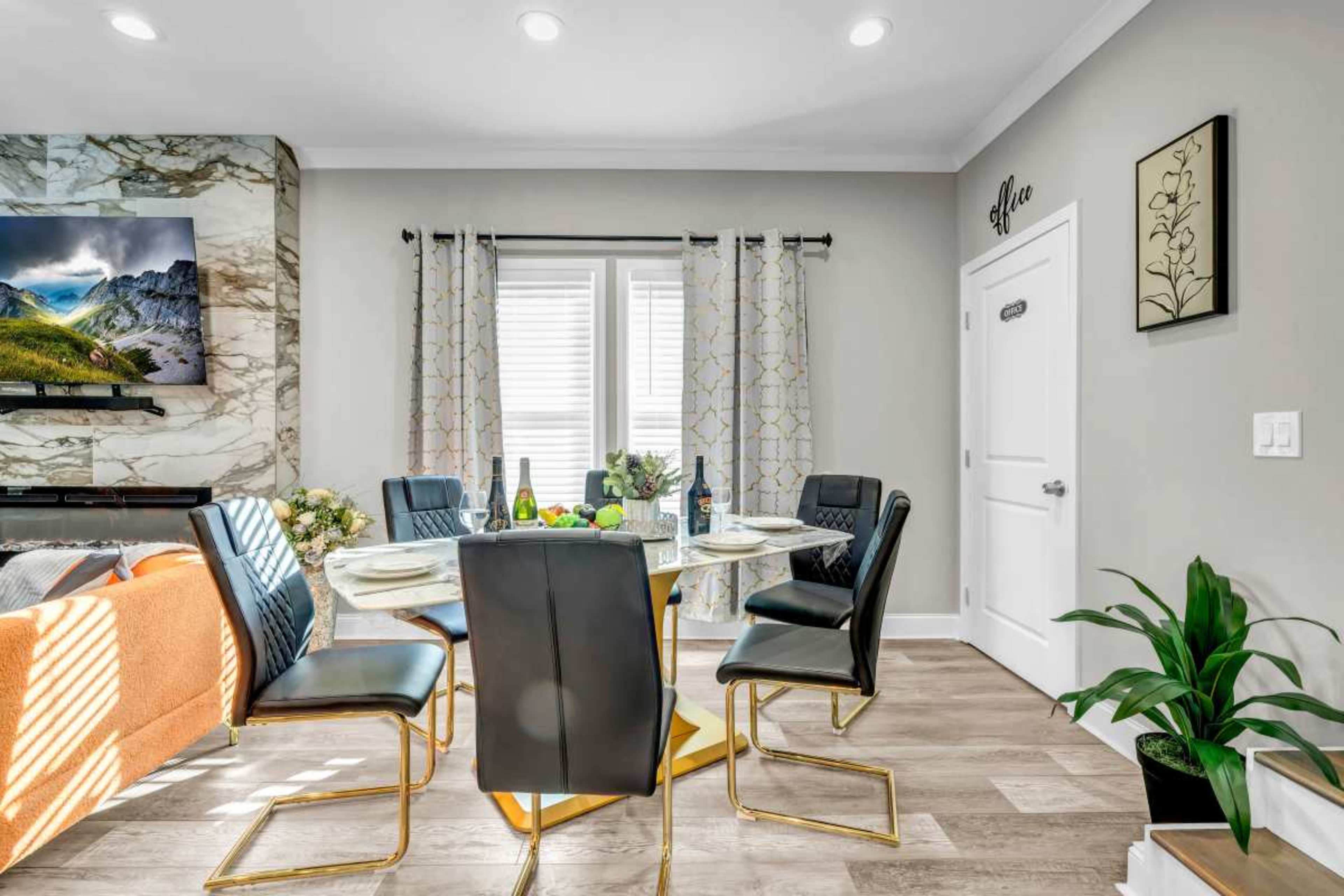 A dining area features a round glass table with black chairs, a light-colored sofa, and a wall-mounted TV above a marble fireplace.