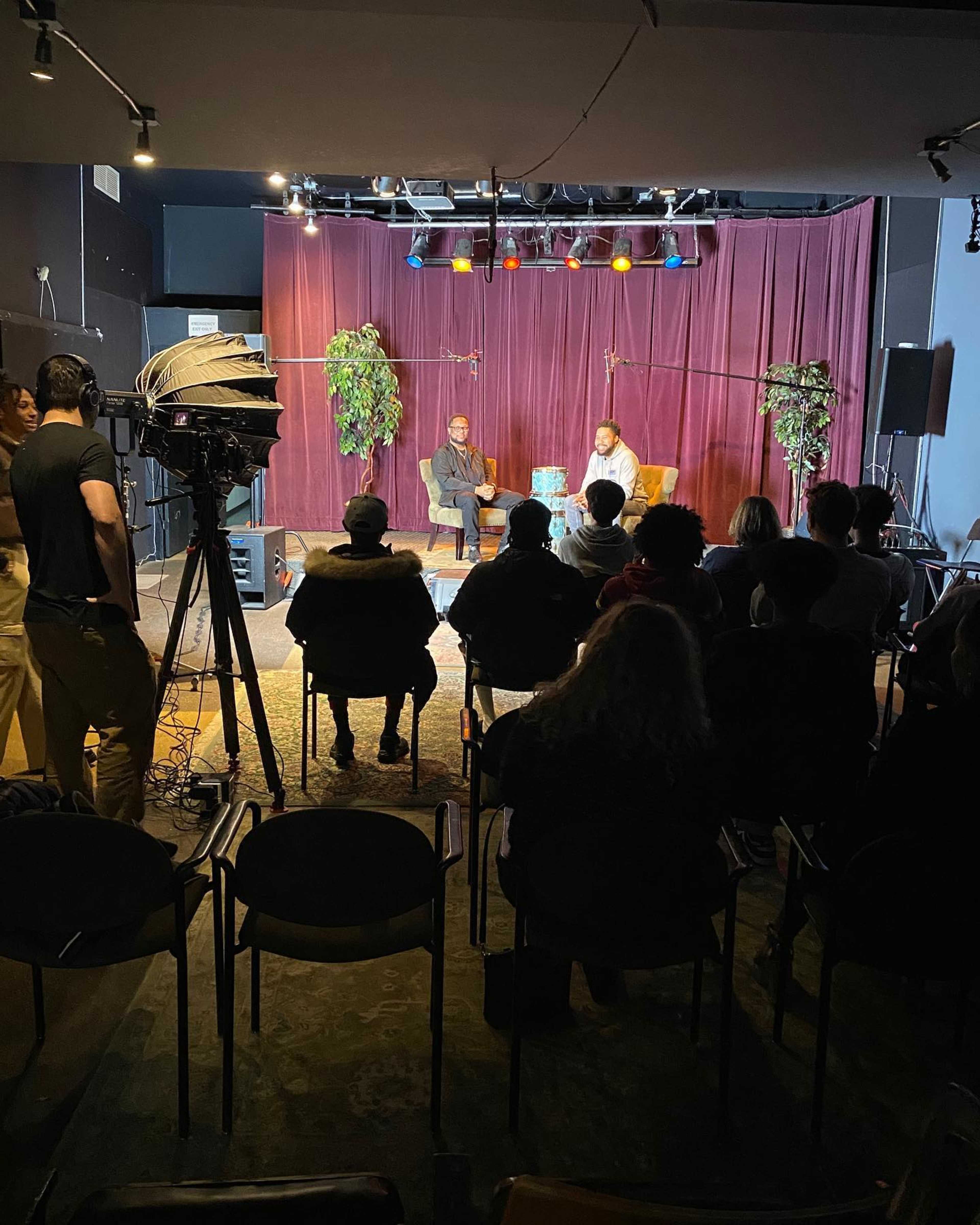 A seated discussion takes place on a stage with two speakers, while an audience watches from rows of chairs and lighting equipment is set up in the background.