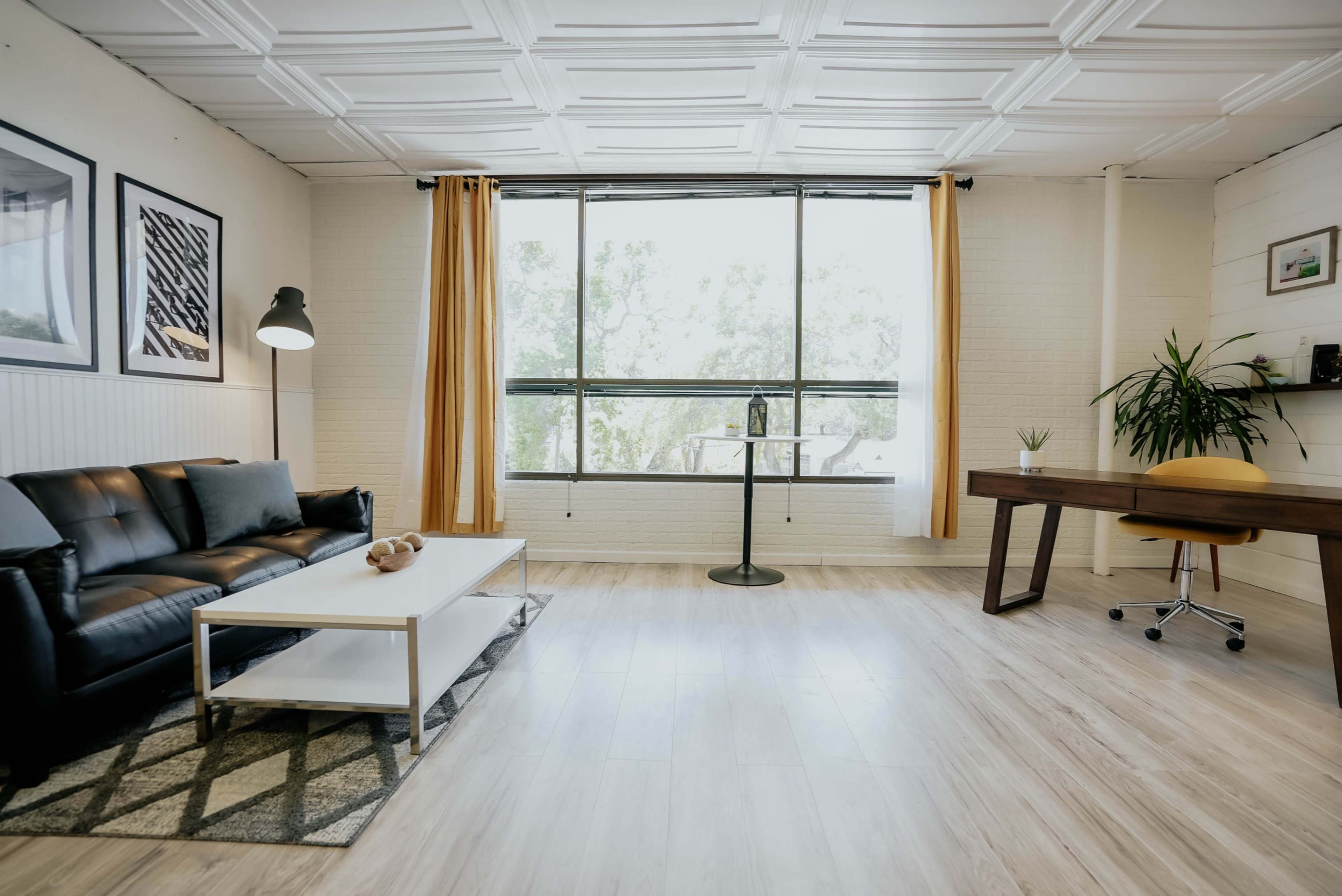 A modern living room features a black sofa, a white coffee table, a yellow curtain, and a desk with a chair, all illuminated by natural light from a large window.