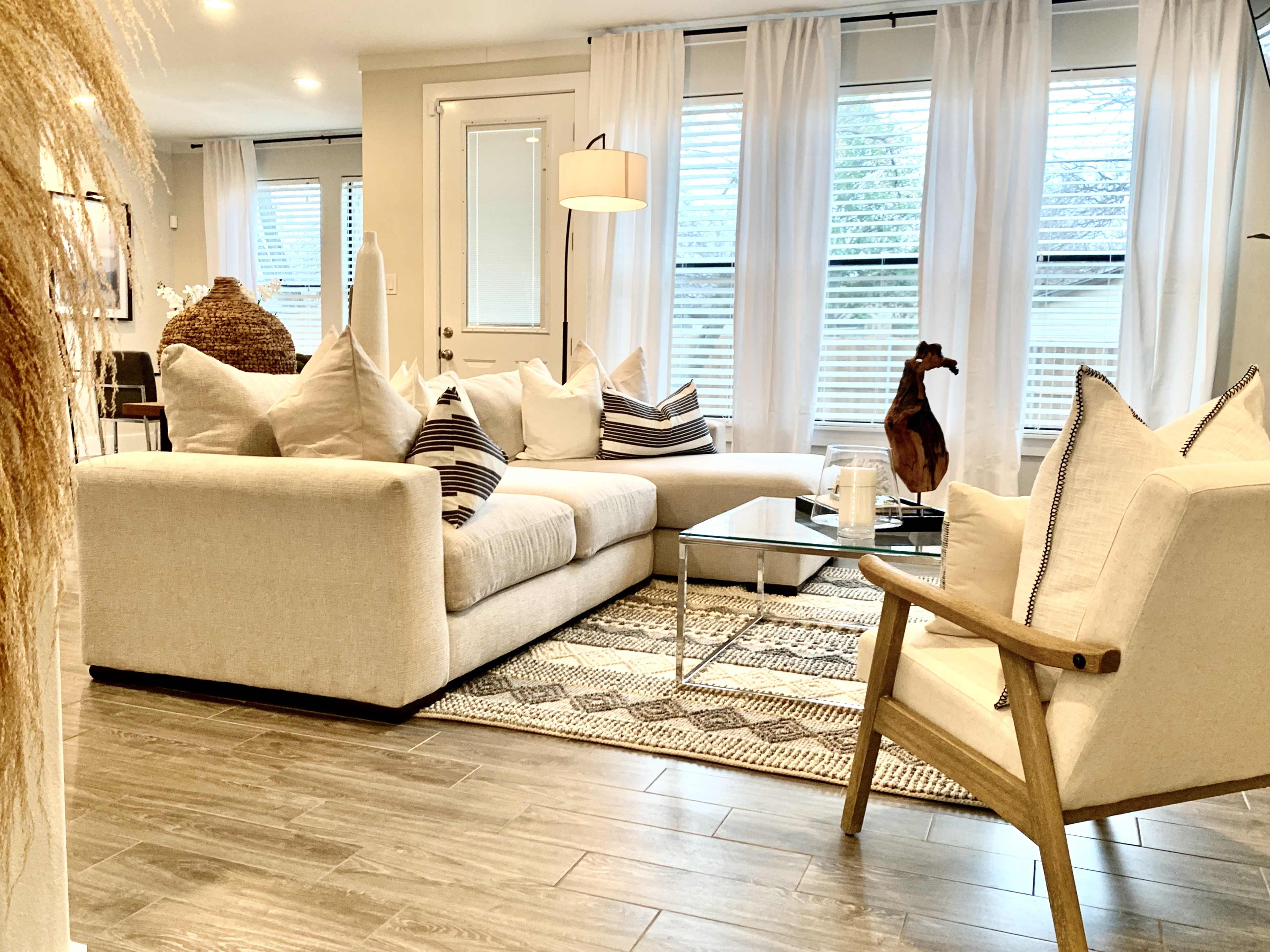 A modern living room features a light-colored sectional sofa, a glass coffee table, and large windows adorned with sheer curtains.