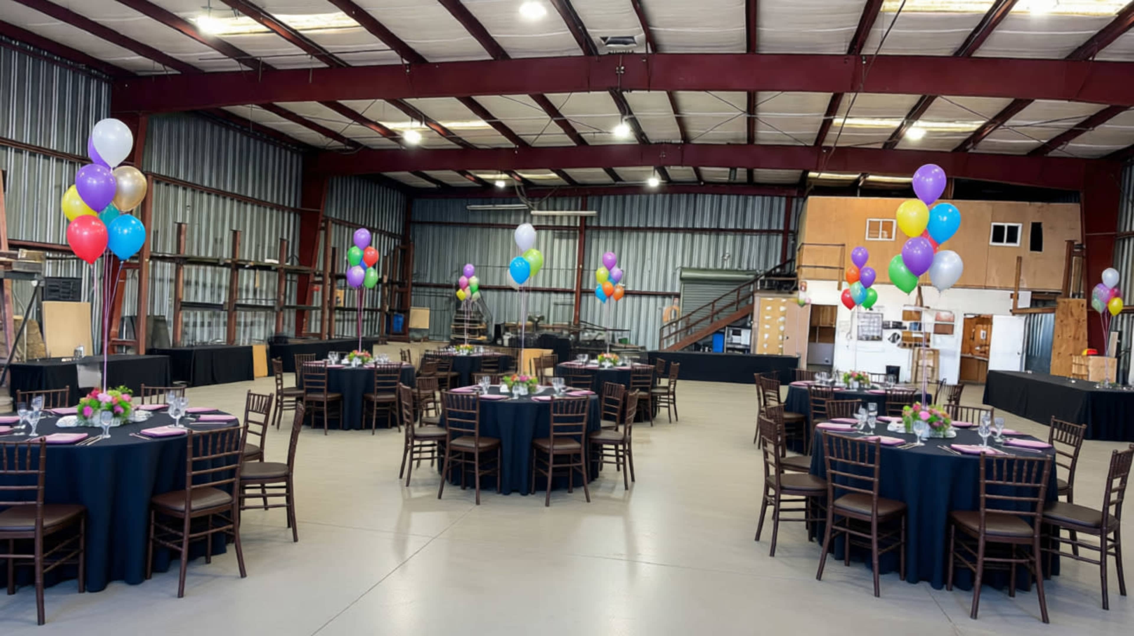 The image shows an indoor event space with several round tables set with tableware and decorated with colorful balloons.