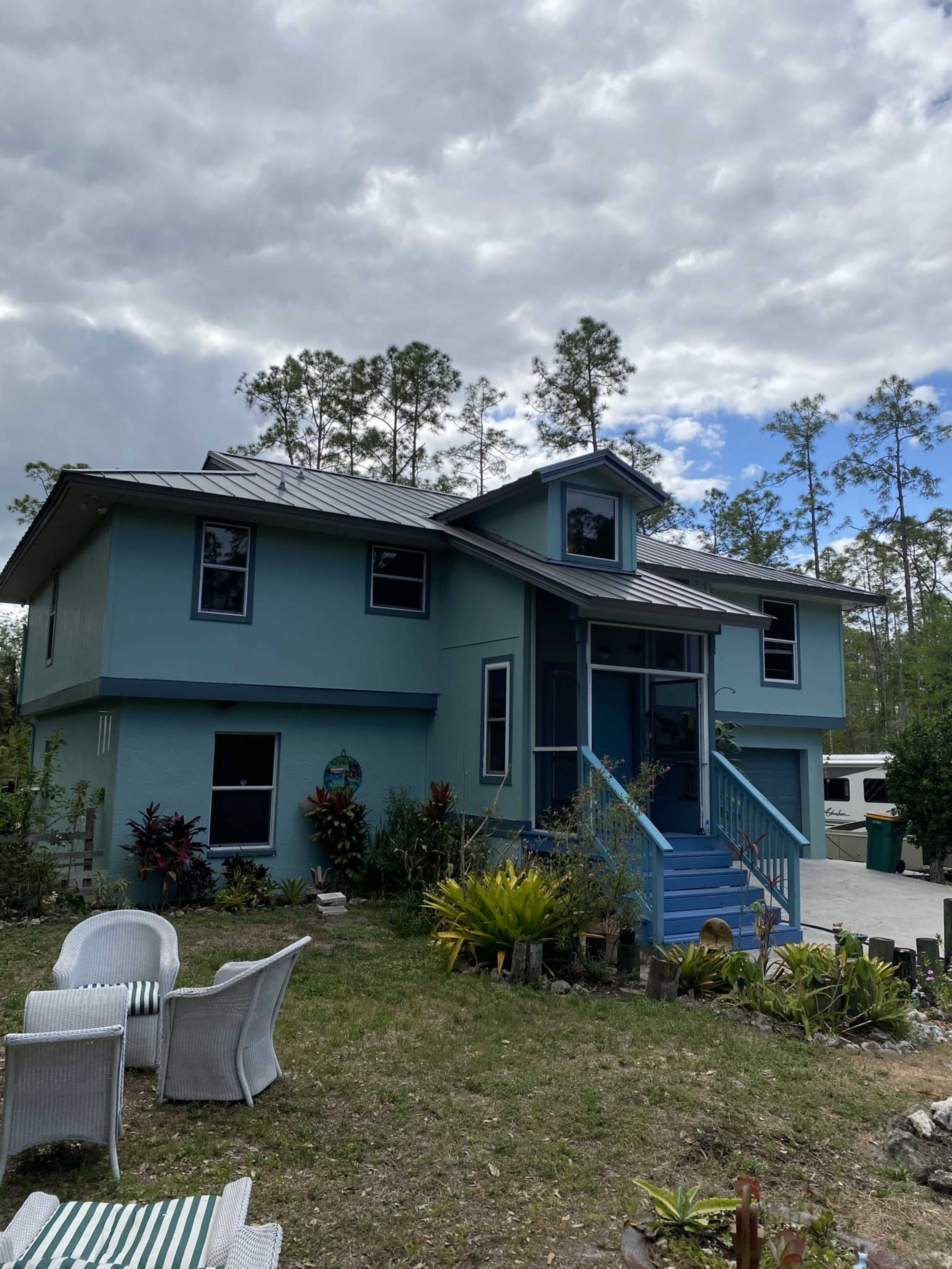 A two-story blue house with a metal roof and a front porch surrounded by greenery and decorative plants.