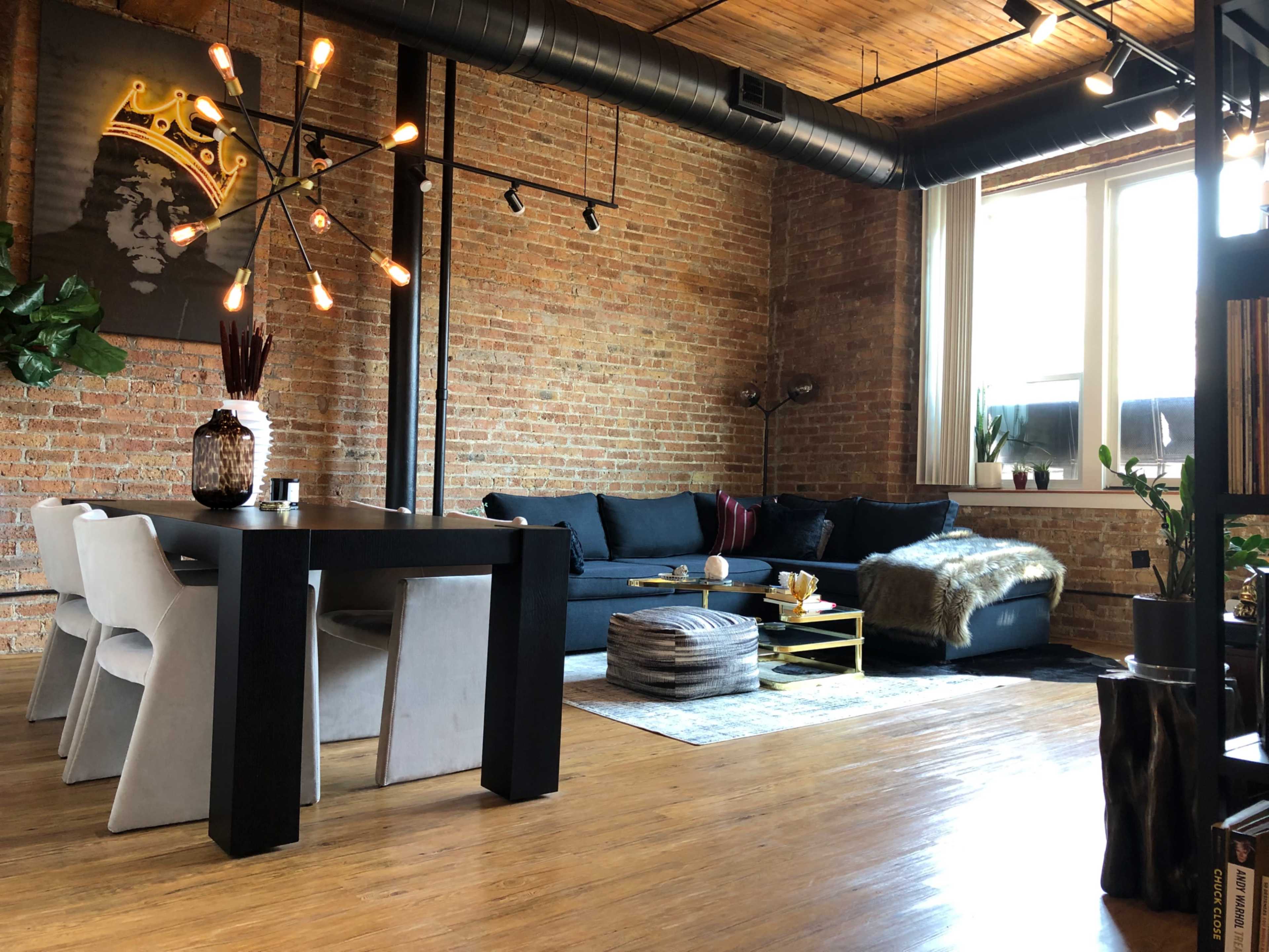 The image shows a modern living space featuring a black sofa, a dining table with gray chairs, a decorative wall art piece, and large windows with natural light illuminating the brick walls.