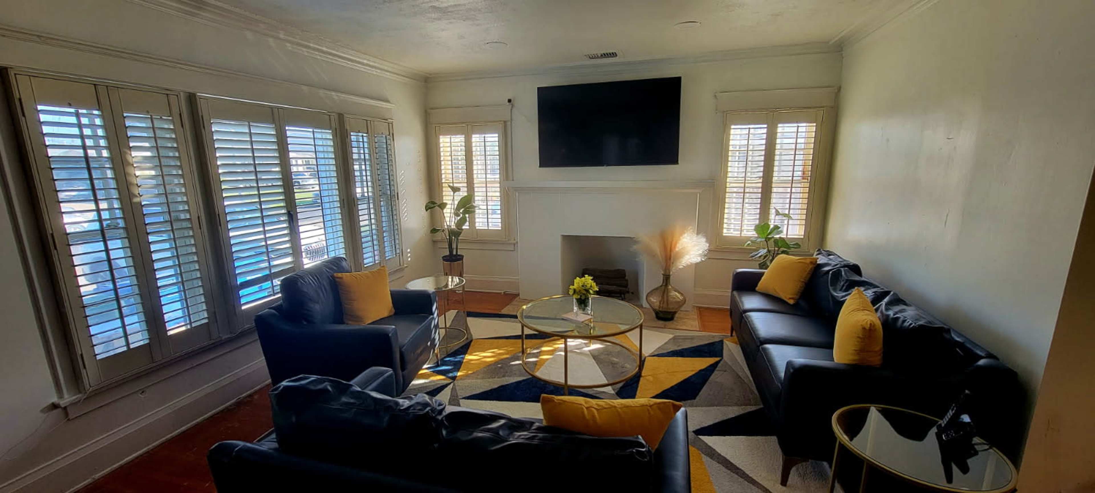 The living room features black leather sofas arranged around a glass coffee table on a geometric rug, with large windows covered by wooden shutters.