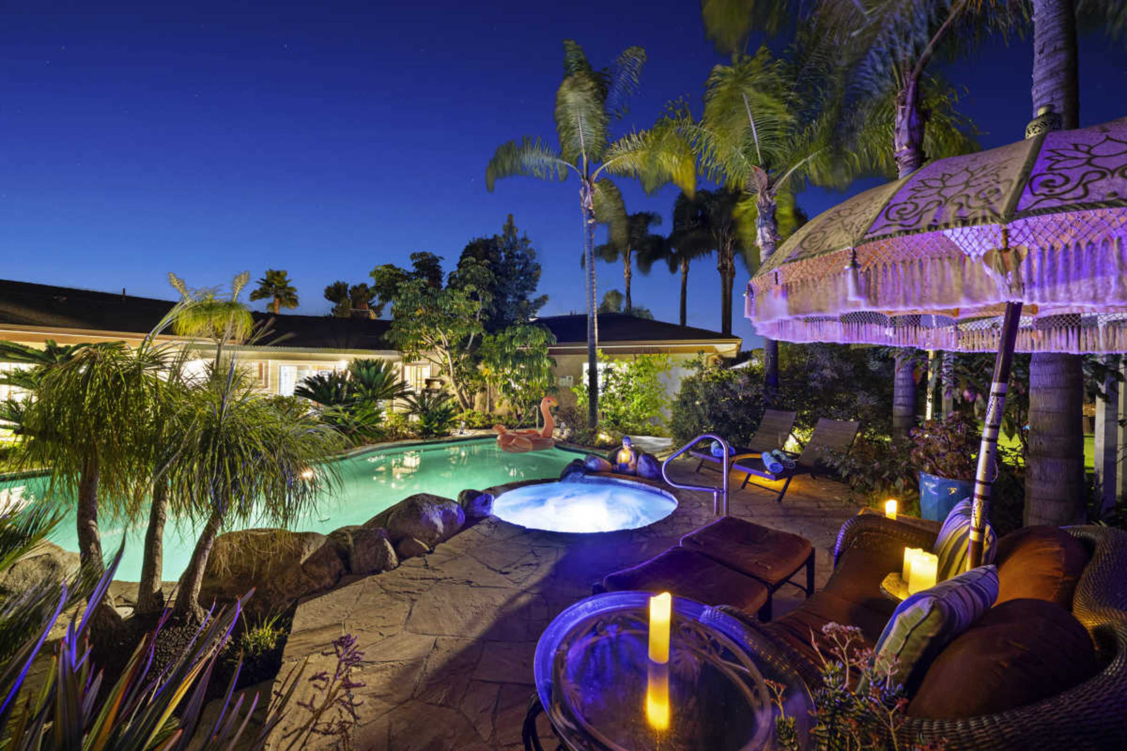 The image shows a backyard pool area illuminated at night, featuring a swimming pool with a spa, surrounded by tropical plants and lounge chairs.
