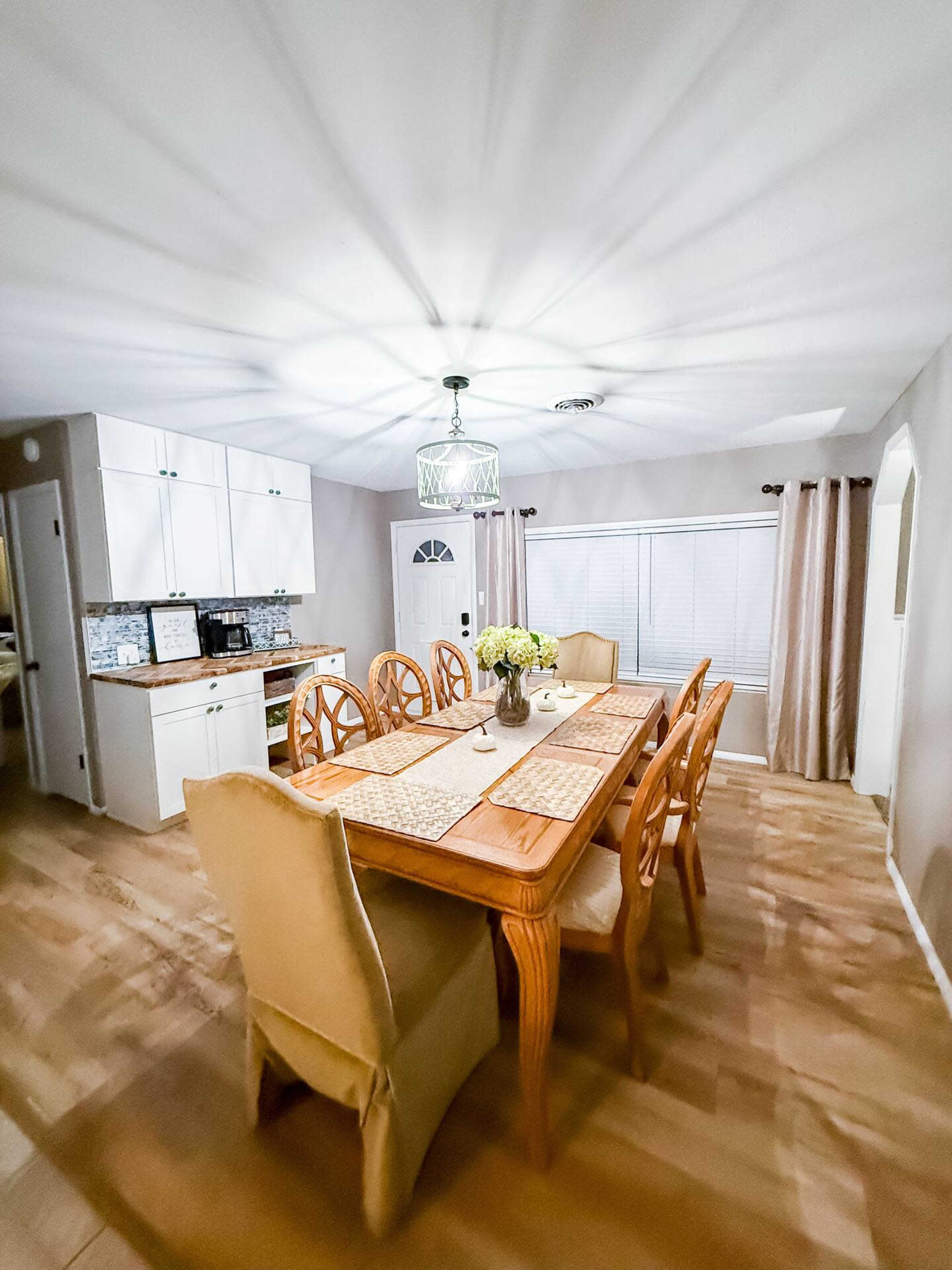 The image shows a dining area with a wooden table surrounded by chairs, a light fixture above, and a kitchen area to the left.