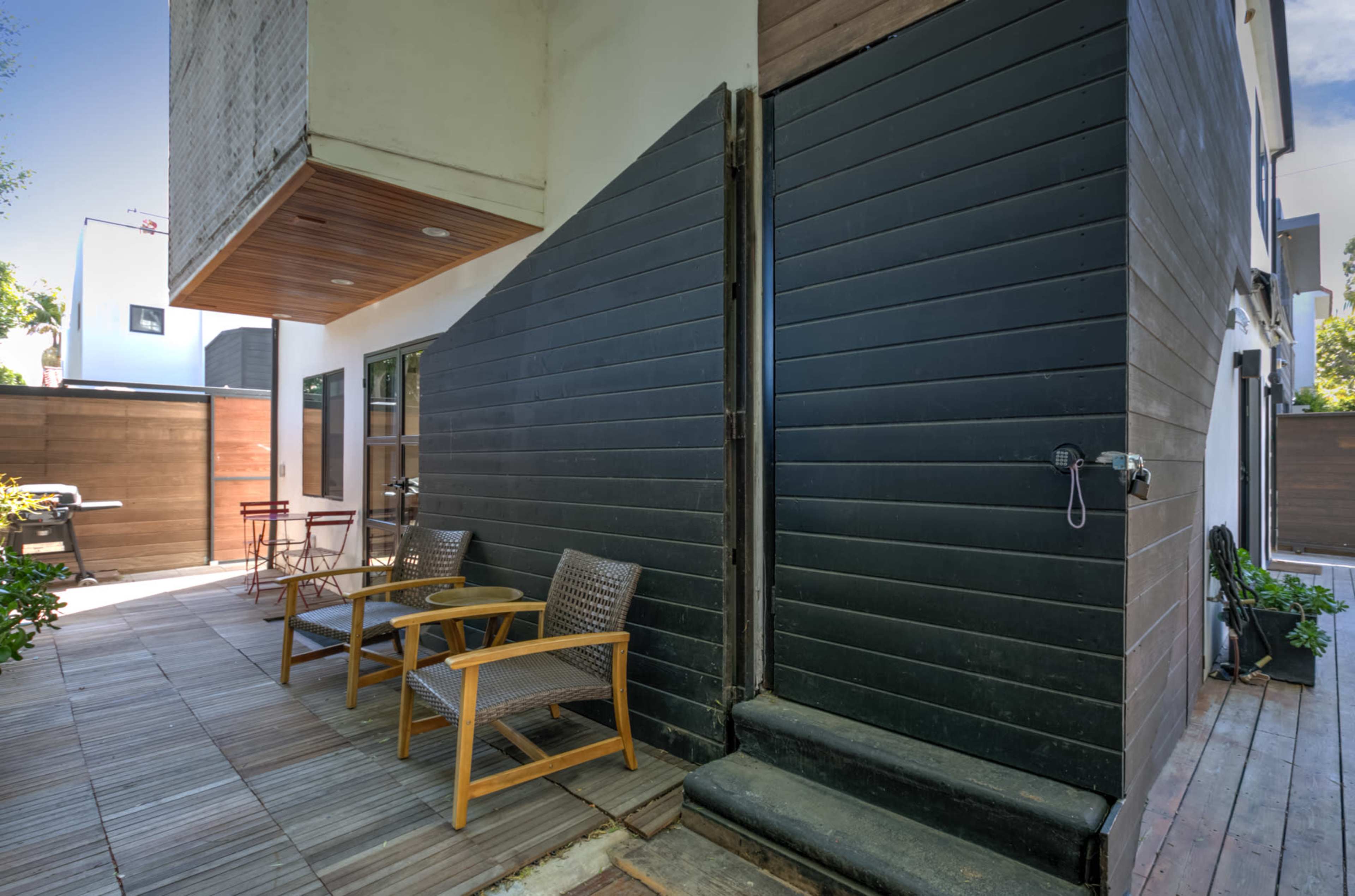 The image shows a modern outdoor patio area featuring two chairs, a wooden deck, and a partially closed door leading into a building.