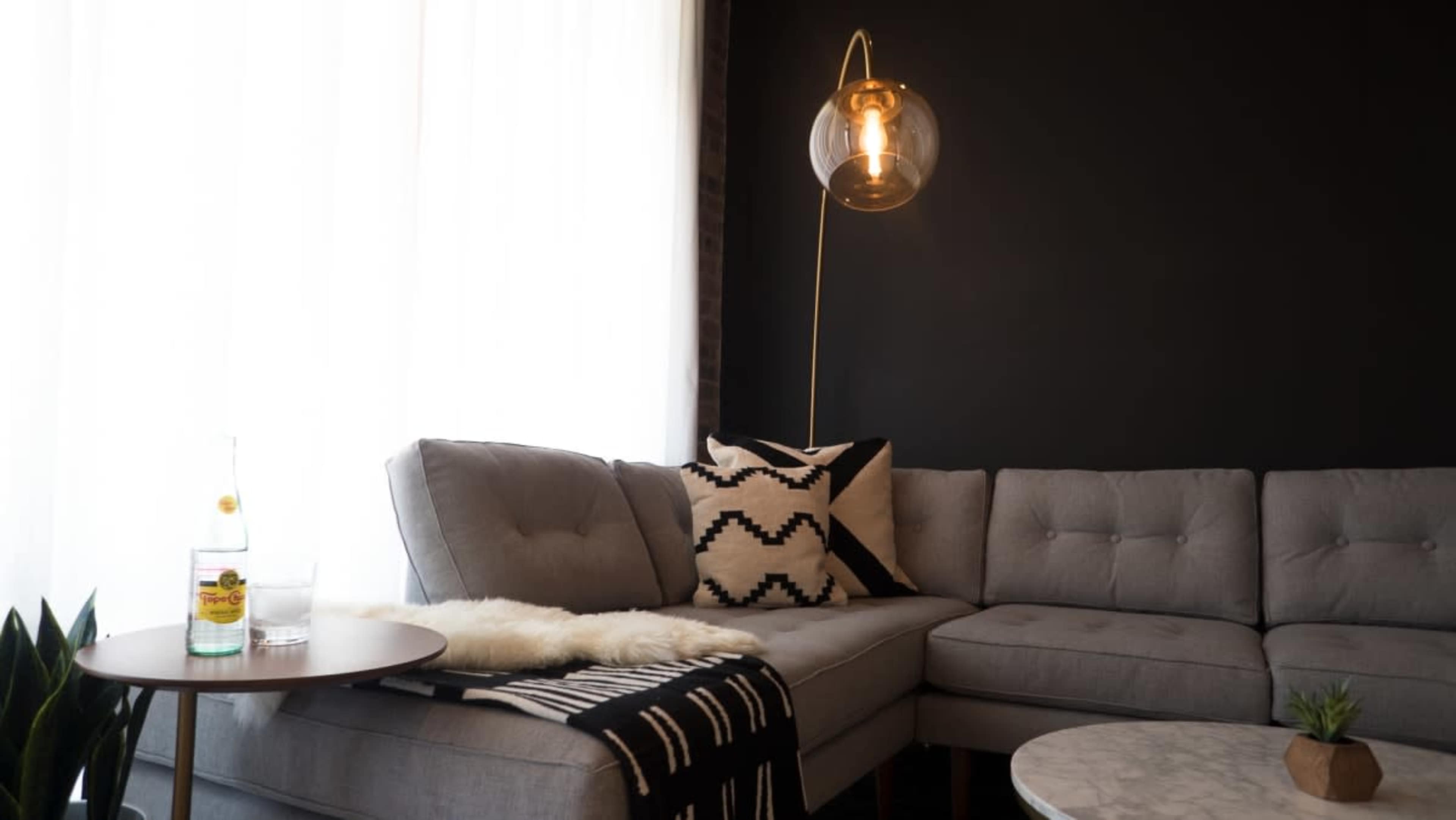 A gray sectional sofa is positioned in a room with dark walls, accompanied by a nearby table with a beverage and a decorative plant.