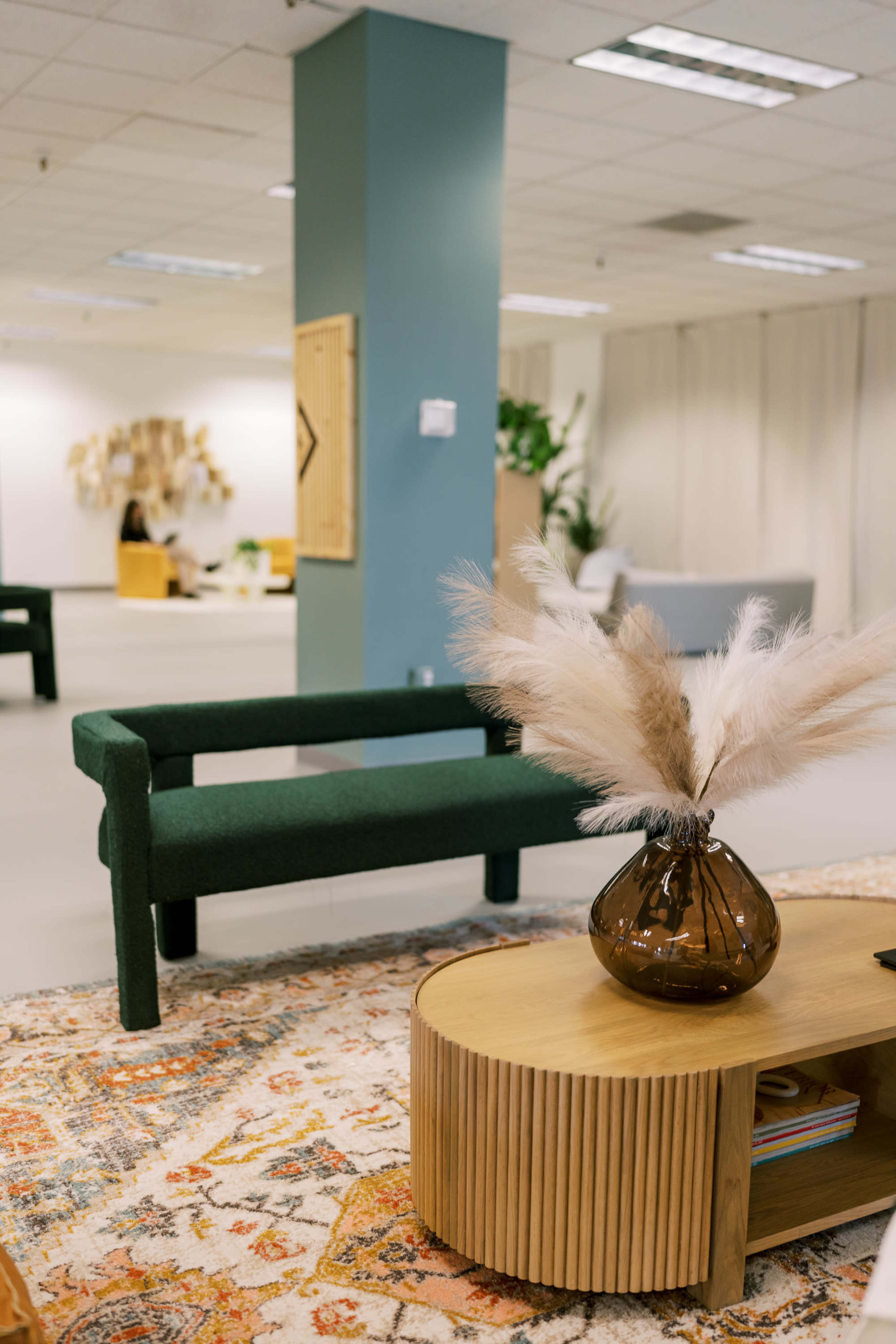 A modern interior space featuring a green sofa and a circular wooden coffee table with a decorative vase and pampas grass, set against a backdrop of neutral-colored walls and furnishings.