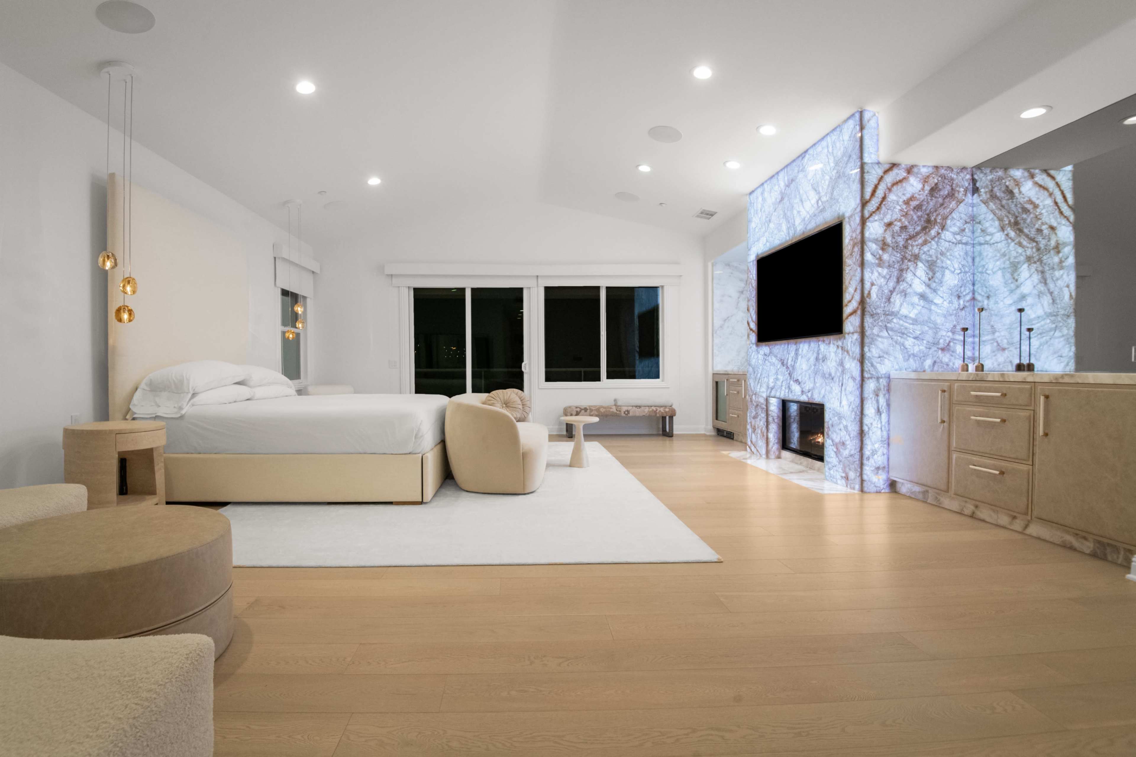 A modern bedroom features a large bed, a seating area, and a striking marble wall with a television, set against large windows.