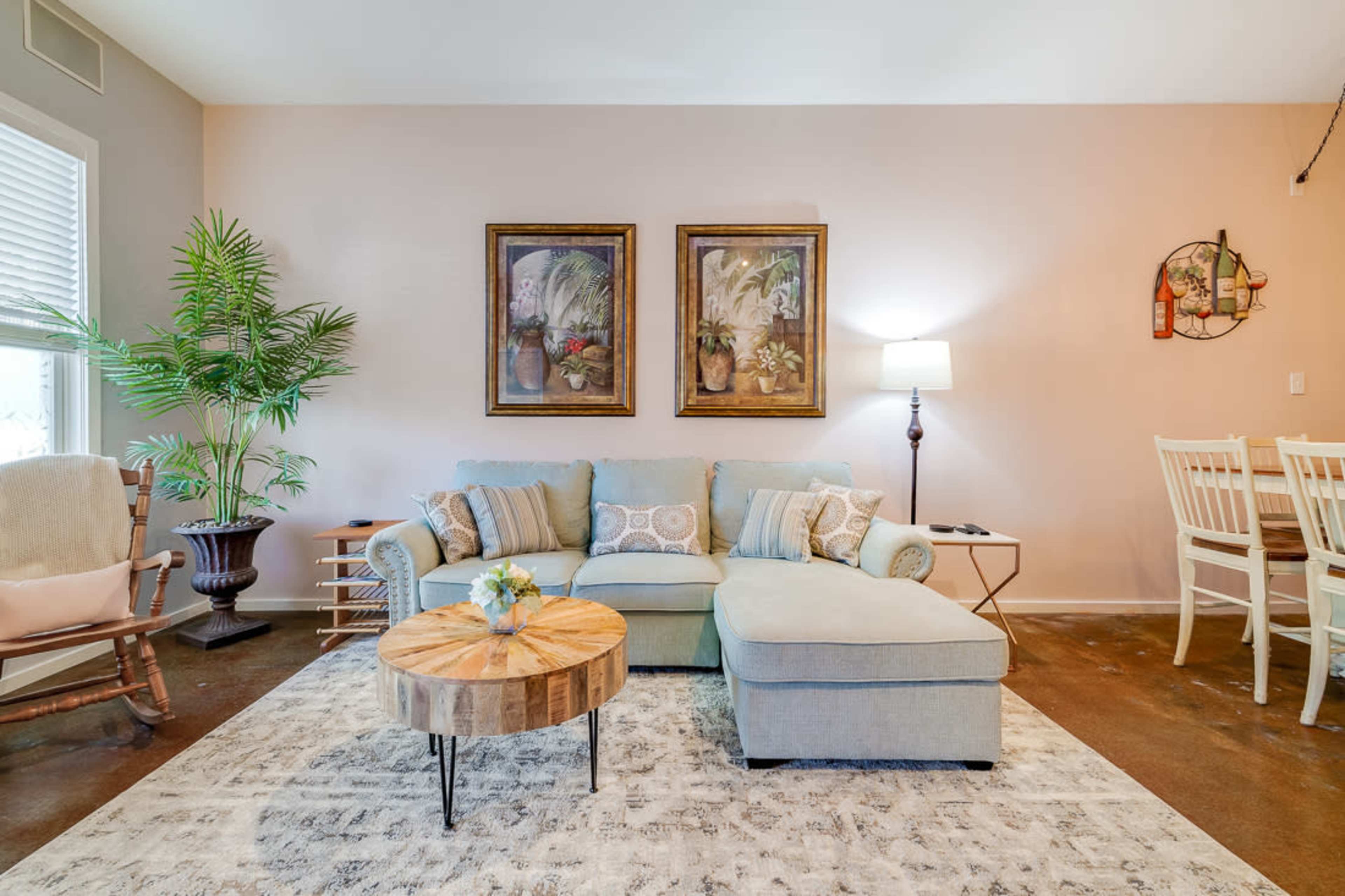 A living room with a light blue sectional sofa, wooden coffee table, two framed paintings on the wall, a standing lamp, and a dining area with white chairs.