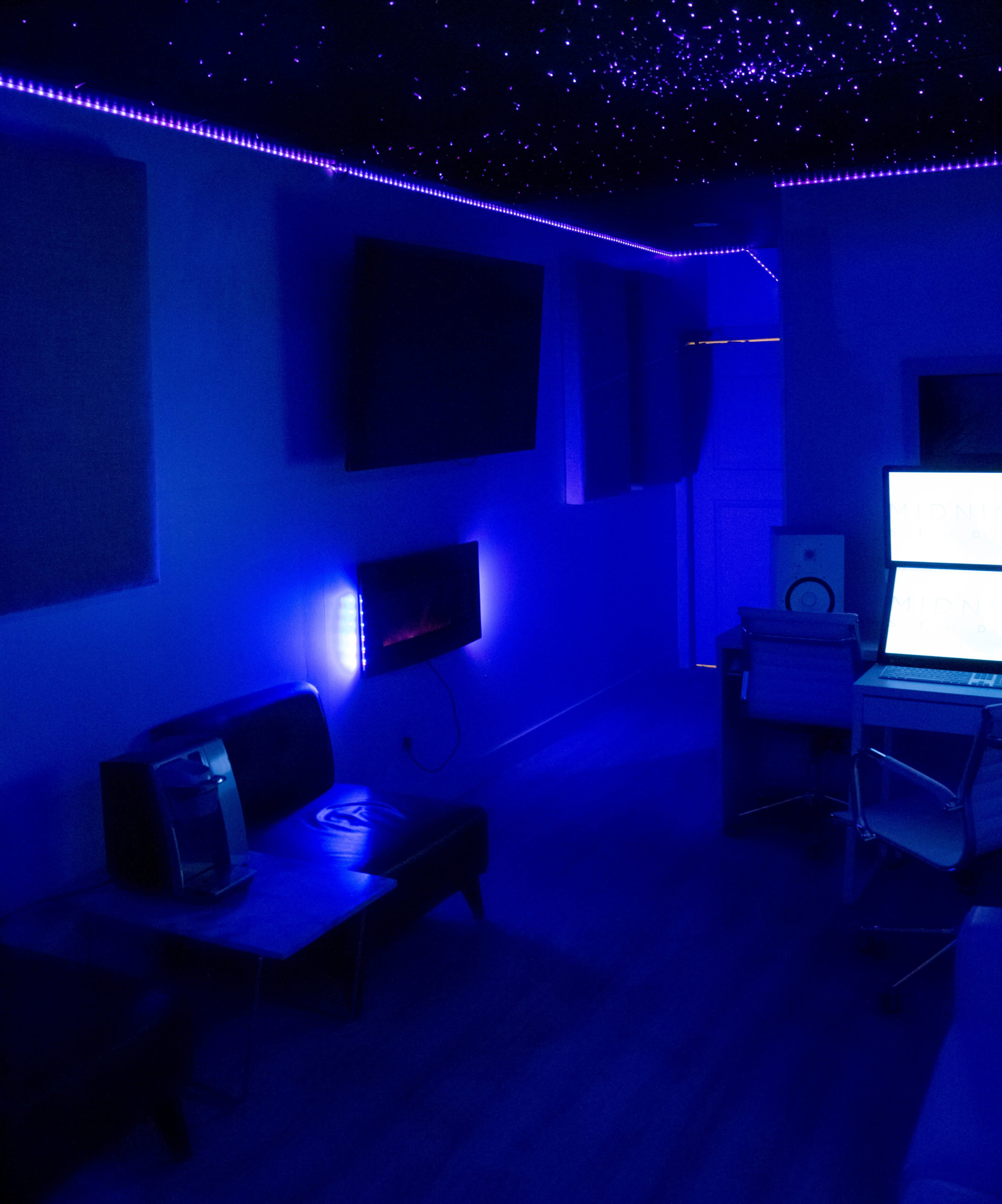 A dimly lit room with blue lighting, featuring a sofa, a television mounted on the wall, and a desk with a computer.