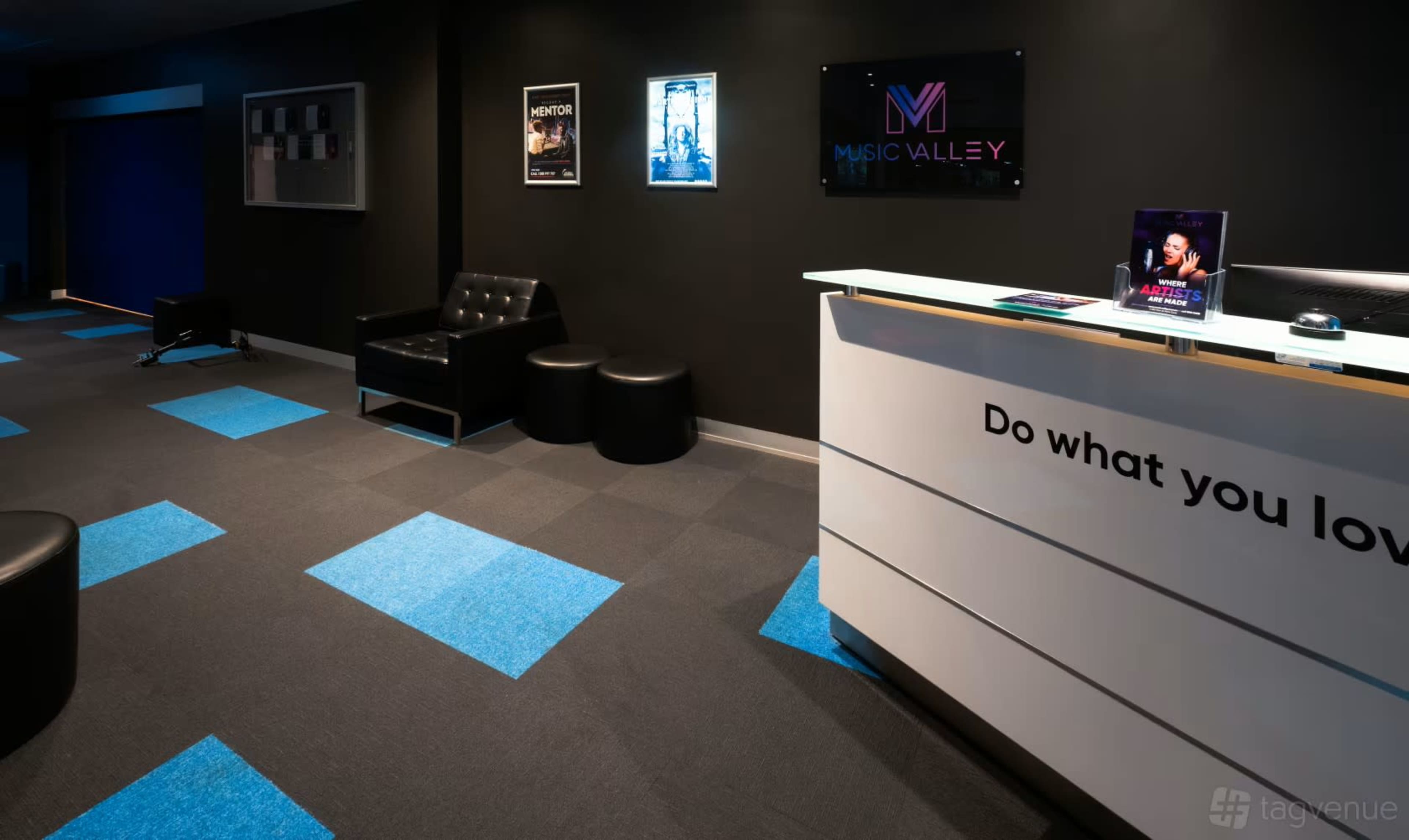 The image shows a modern reception area with a black wall, a sleek white desk featuring the "Music Valley" logo, and colorful blue carpet tiles.