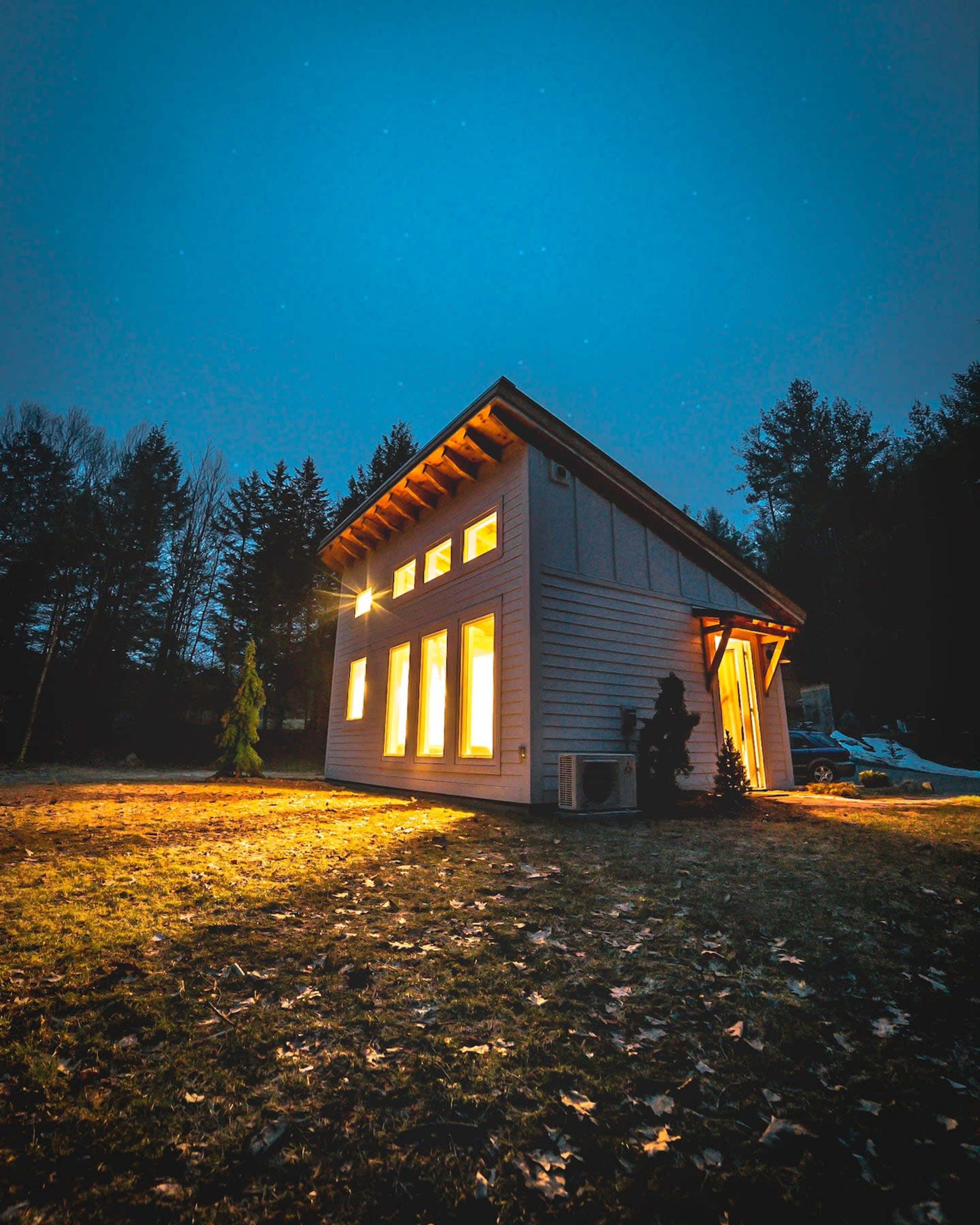 A modern house with large windows is illuminated at dusk, surrounded by trees and a grassy area.