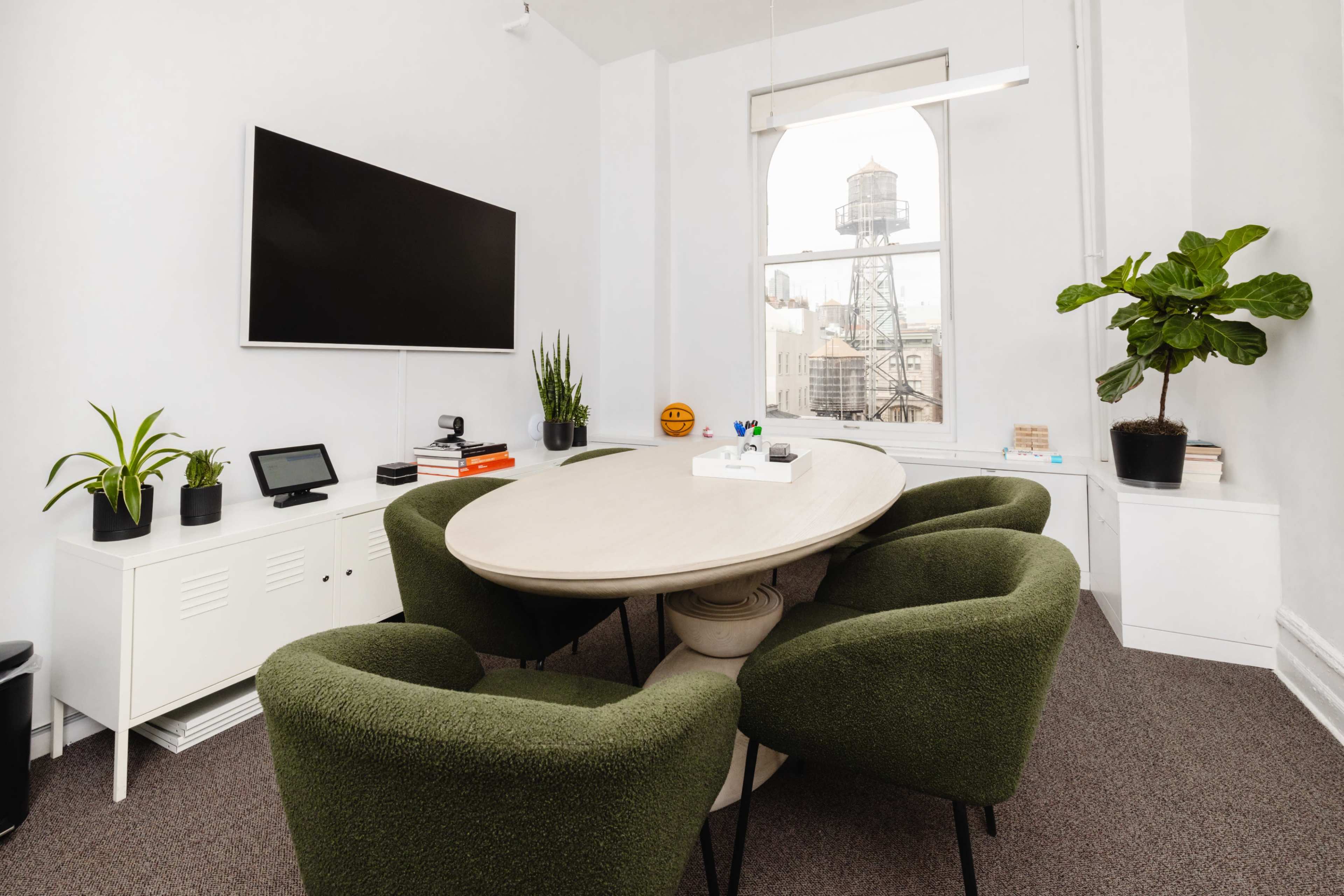 A modern meeting room features a large oval table surrounded by green chairs, a wall-mounted television, and potted plants, with a view of a water tower through the window.
