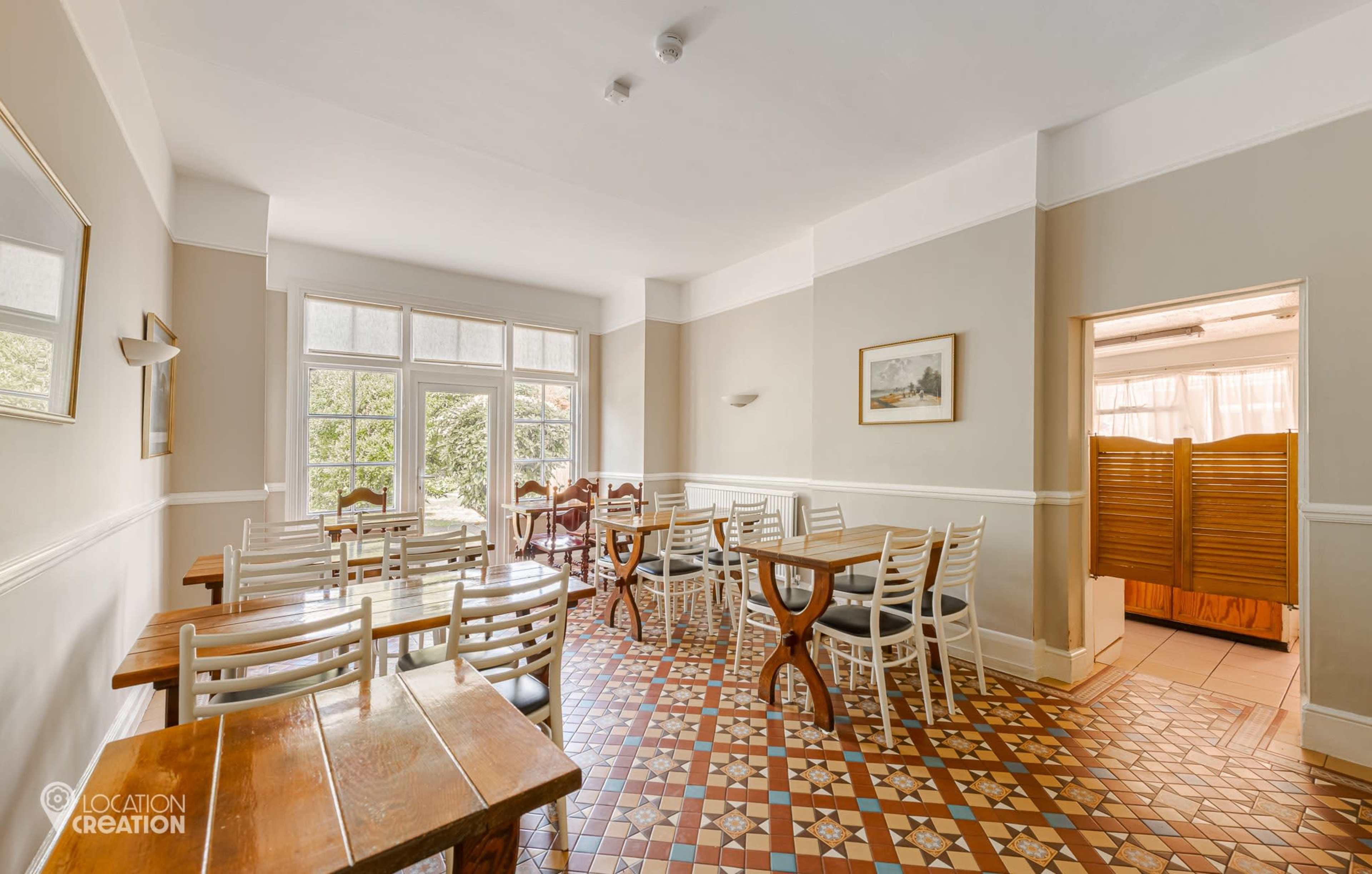 The image shows a dining area with wooden tables and white chairs, featuring tiled flooring and large windows that let in natural light.