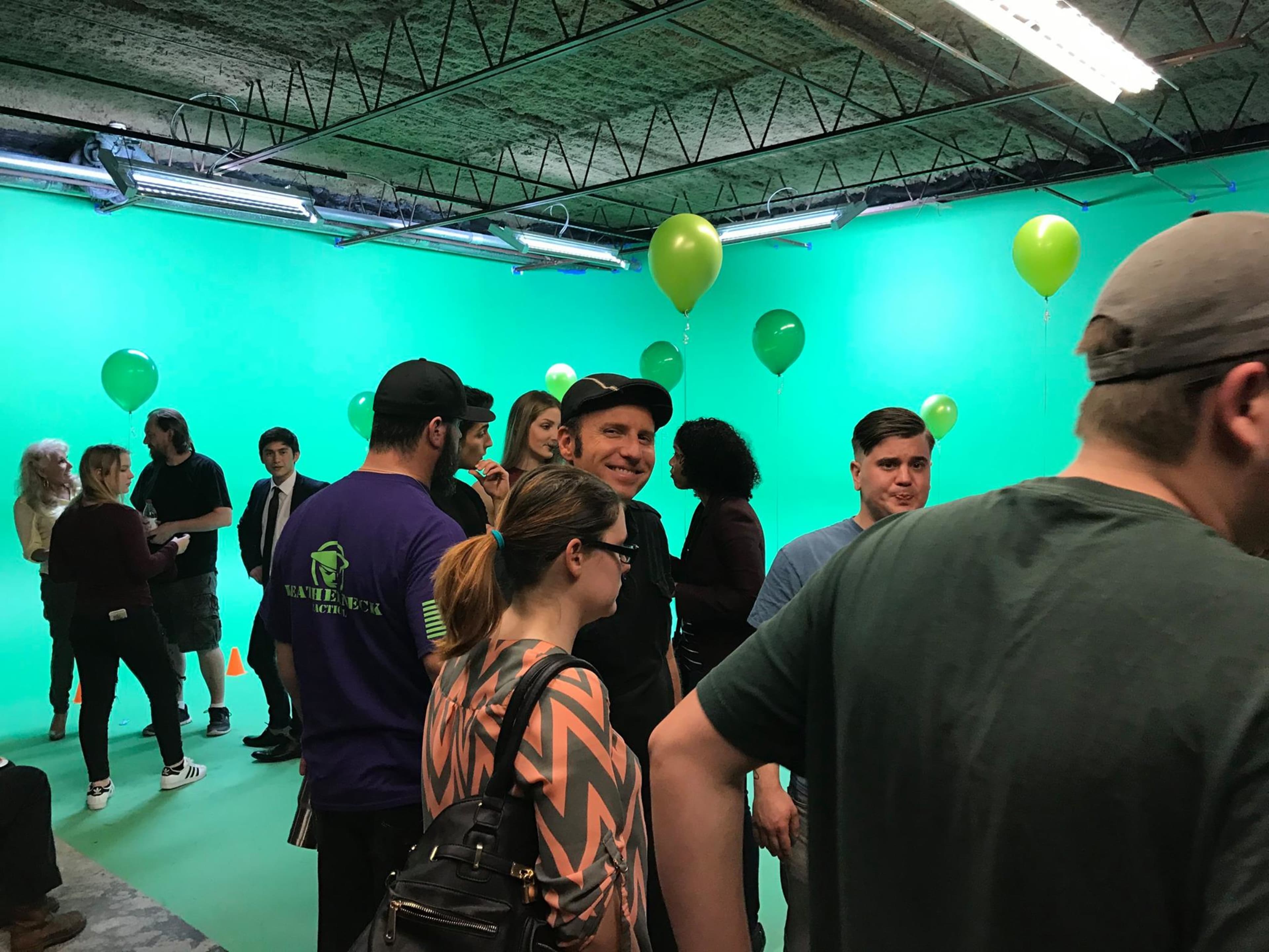 A group of people mingles in a studio with a green backdrop and green balloons.