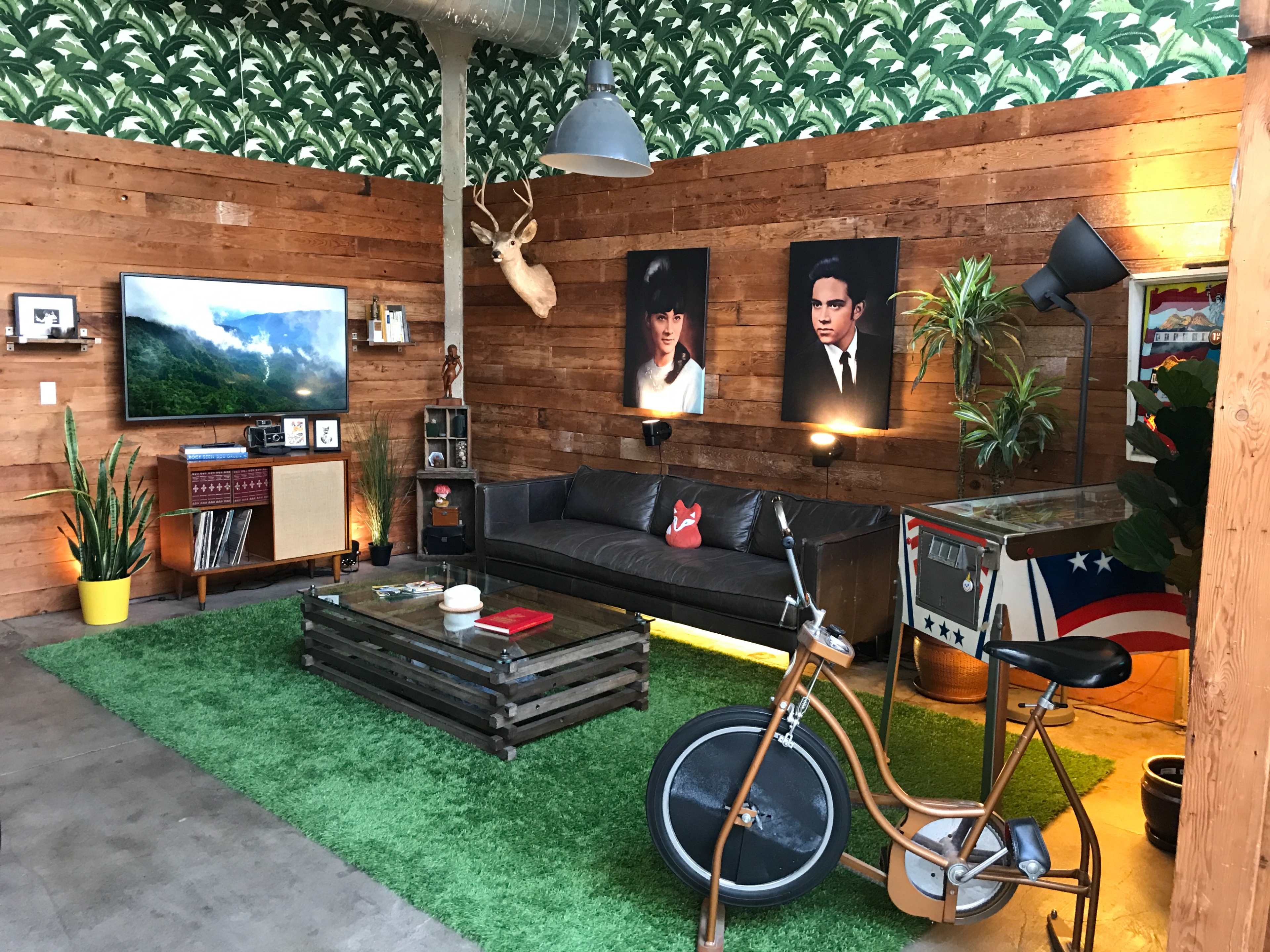 A stylish living room with wooden walls, a large TV, a black sofa, a green rug, and decorative plants, featuring artwork and a vintage bicycle.