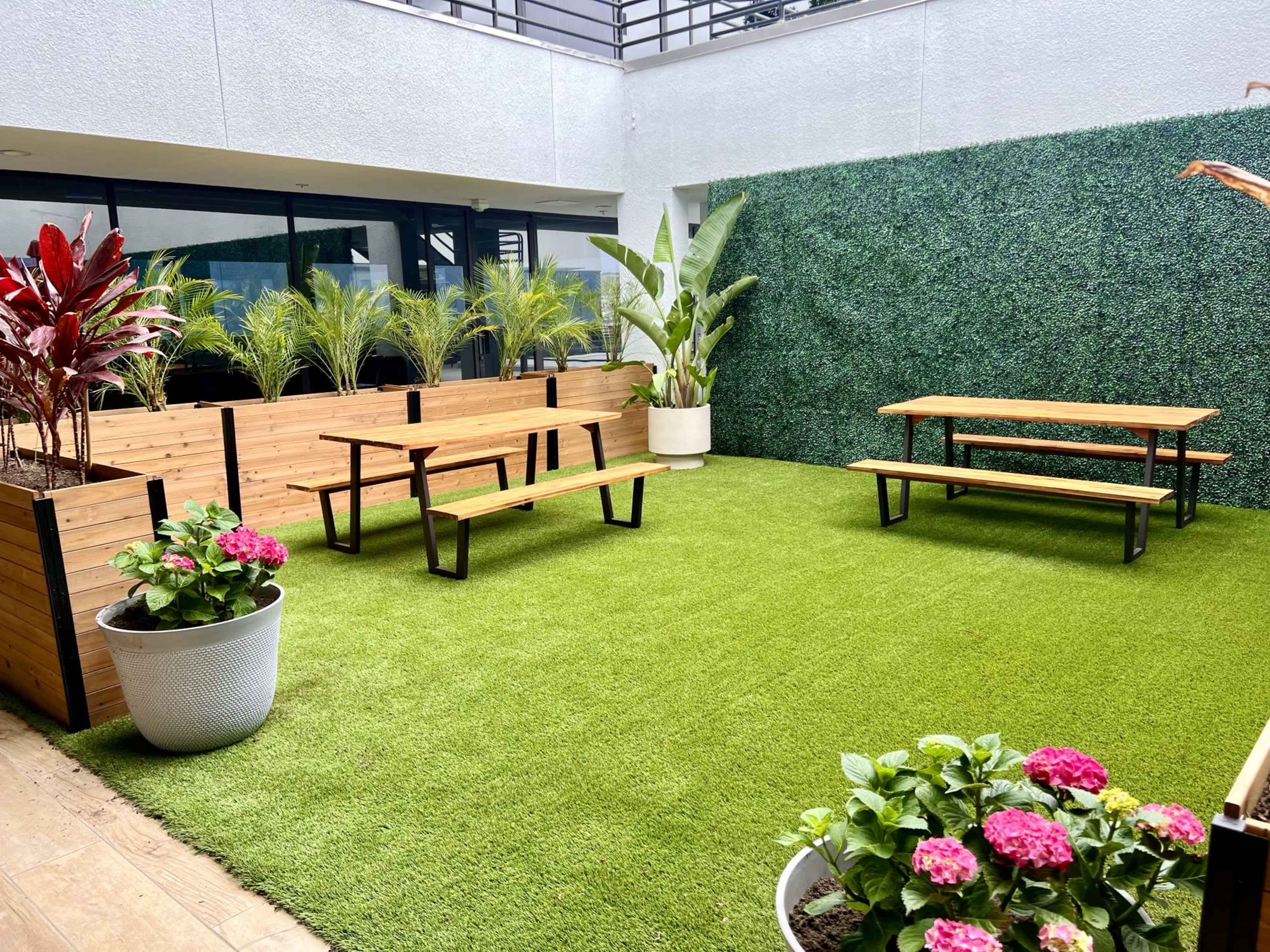 A green lawn area enclosed by wooden planters, featuring two wooden picnic tables and potted plants with pink flowers.