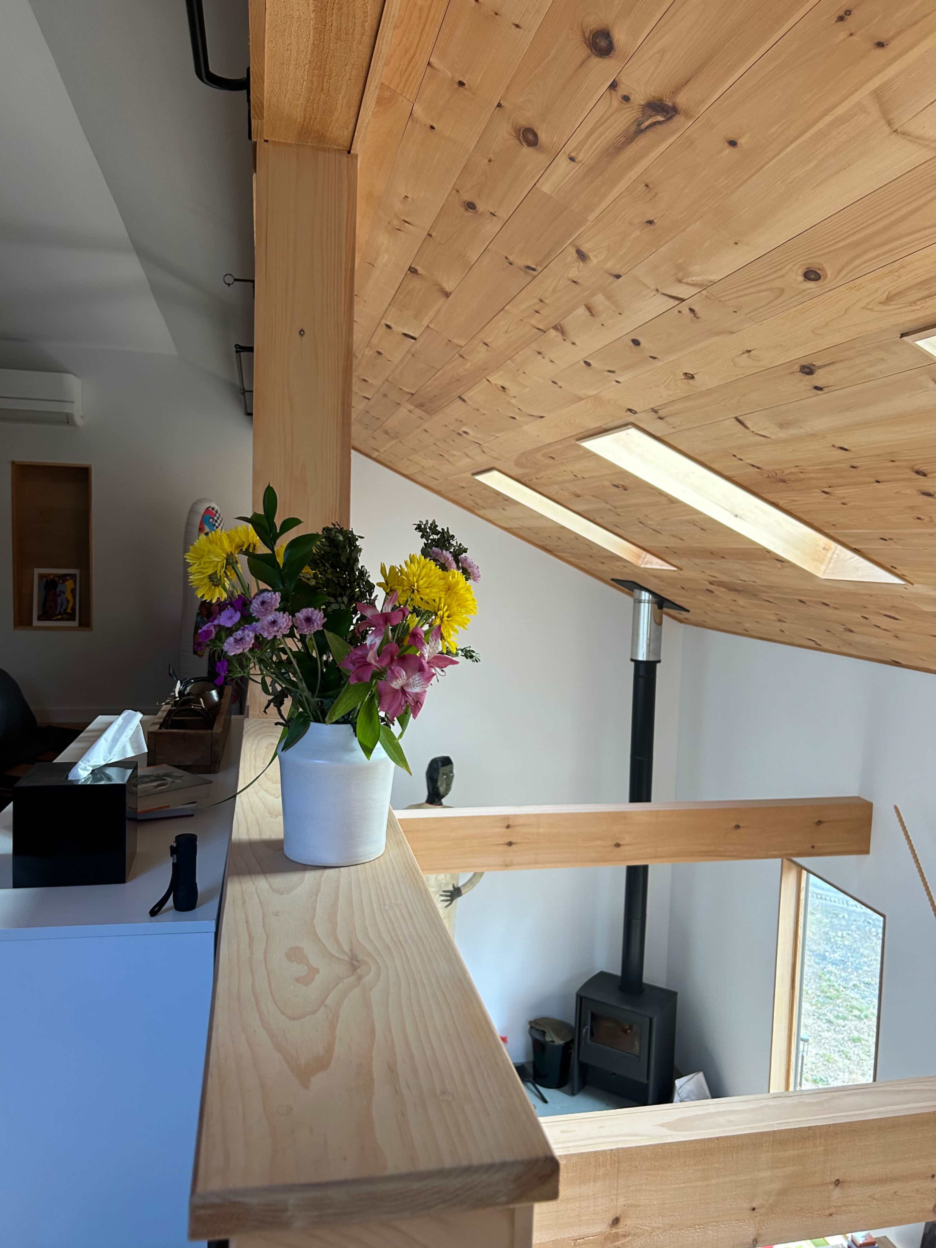 A vase of flowers sits on a wooden ledge in a room with a sloped ceiling and a view of a wood stove below.