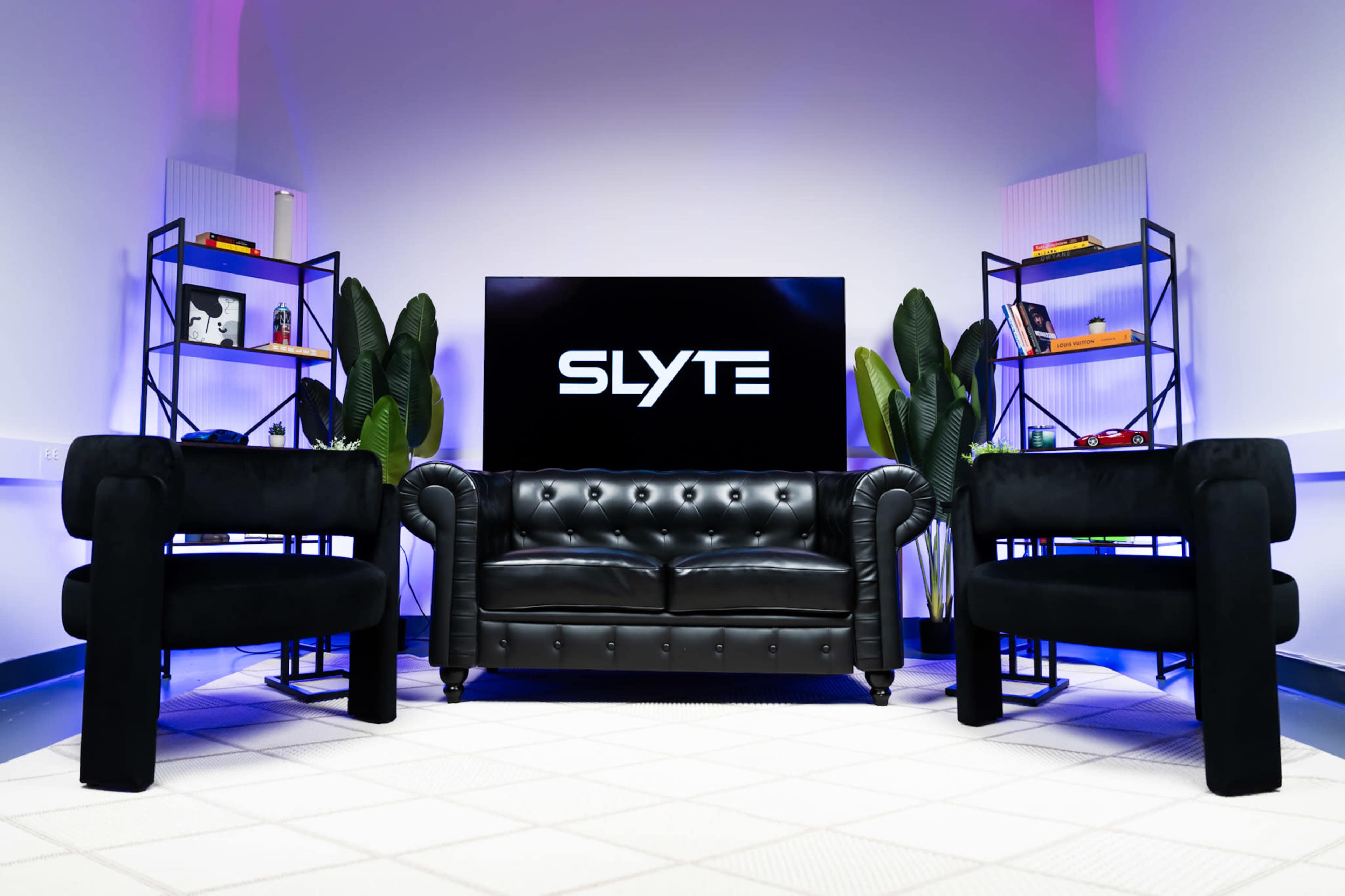 The image shows a modern living space featuring a black leather chesterfield sofa, two black armchairs, and shelving units adorned with decorative items, all in front of a large screen that displays the word "SLYTE."