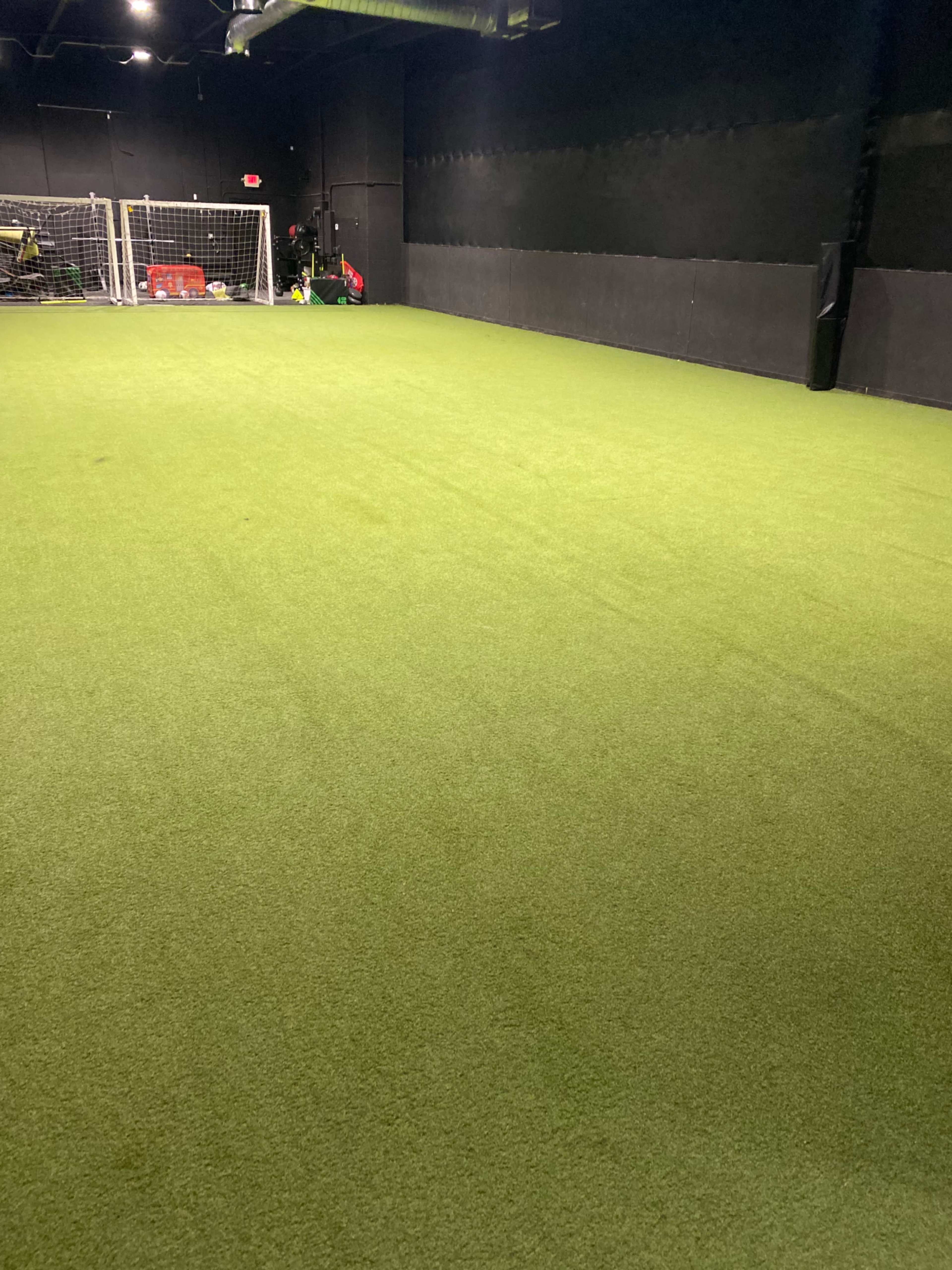 The image shows an indoor training facility with artificial turf and a soccer goal at one end.