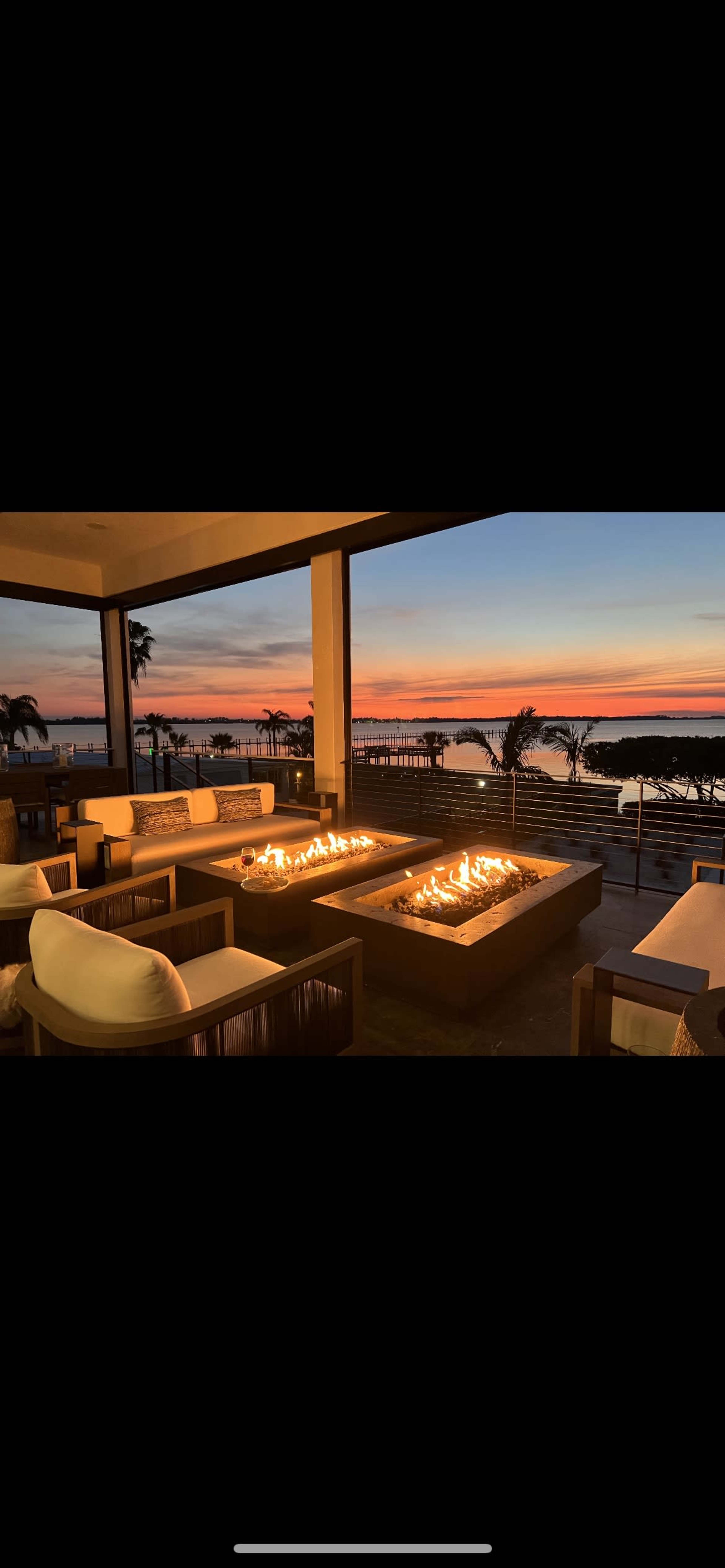A modern outdoor lounge featuring fire pits, comfortable seating, and a view of a sunset over water.