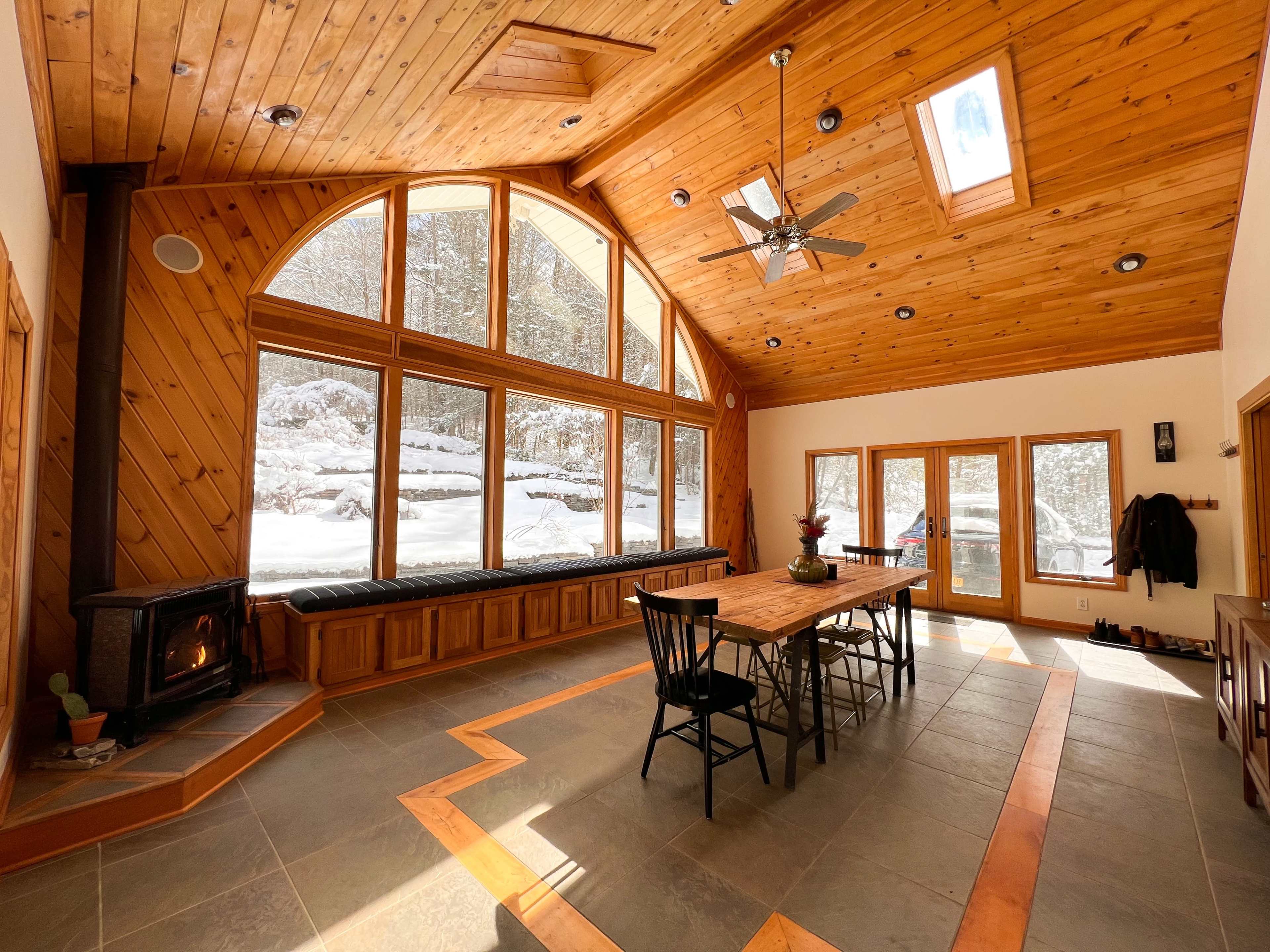A spacious wooden interior with large windows overlooking a snowy landscape, featuring a dining table at the center and a wood stove in the corner.