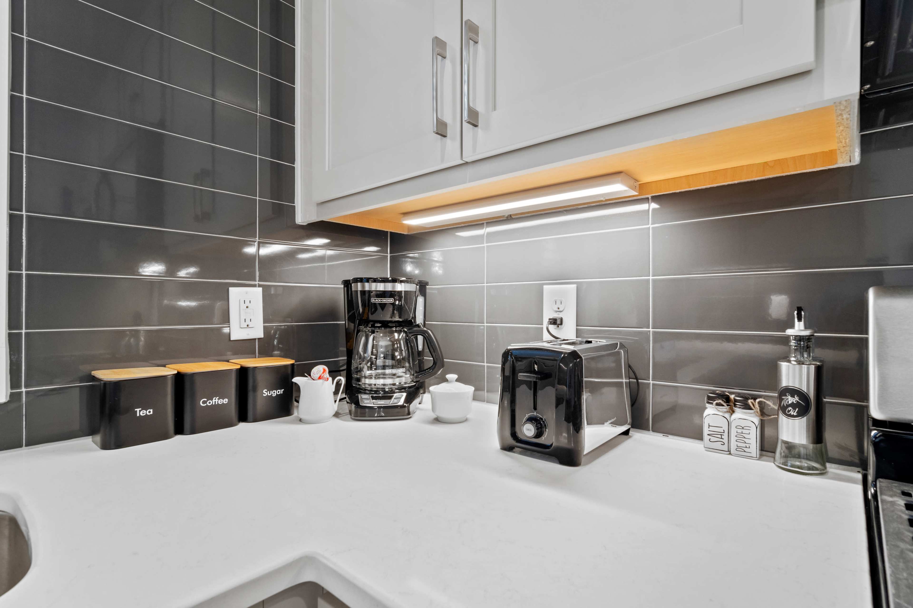 A modern kitchen countertop features a coffee maker, toaster, and containers labeled for tea, coffee, and sugar, with a backsplash of glossy gray tiles.